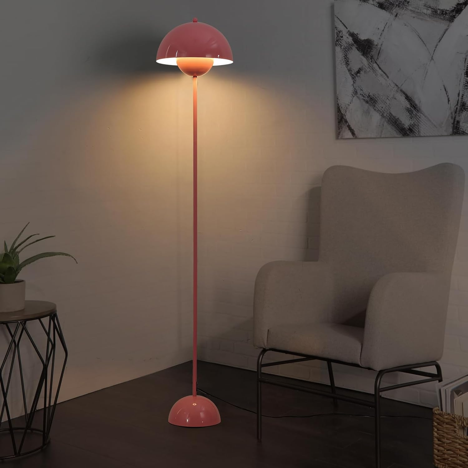 Una lámpara de pie rosa con una pantalla redonda, iluminando una habitación oscura con una mesa auxiliar y una pintura.