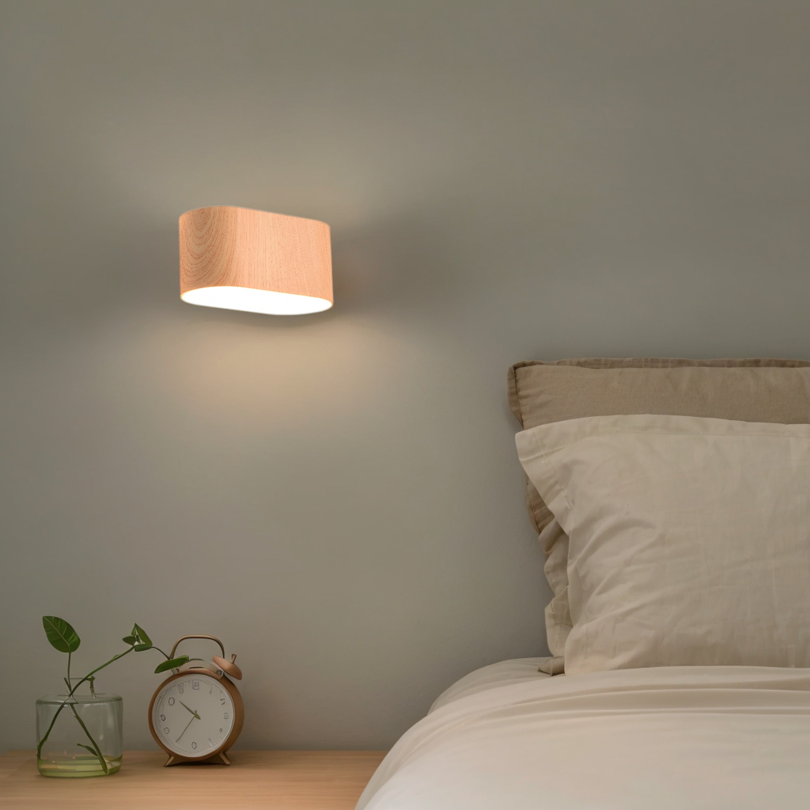 Lámpara de pared de madera sobre una cama. Un reloj y una planta en la mesita de noche. Ropa de cama beige.
