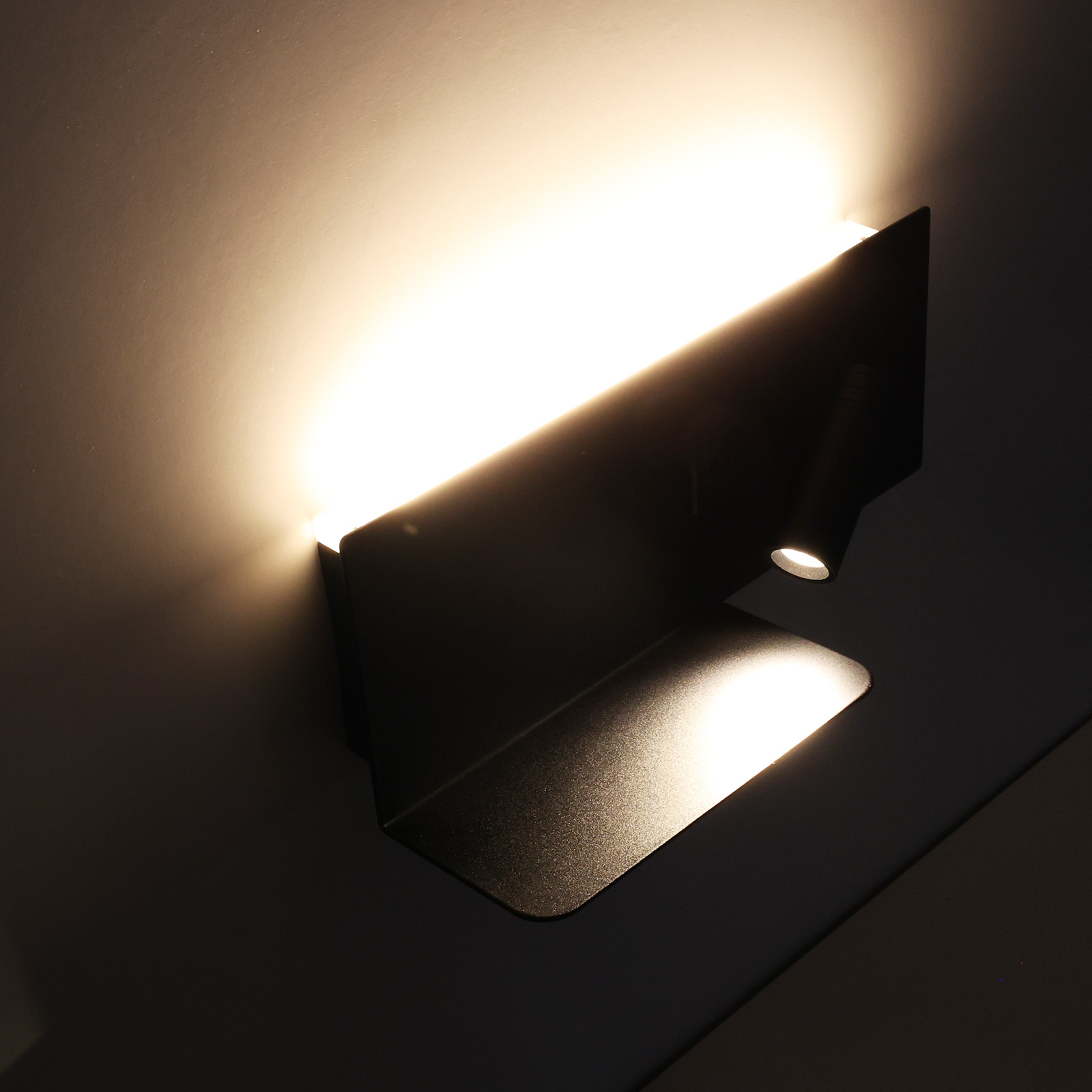 Lámpara de pared negra, con un pequeño foco y una fuente de luz más grande arriba. Luz brillante brillando.