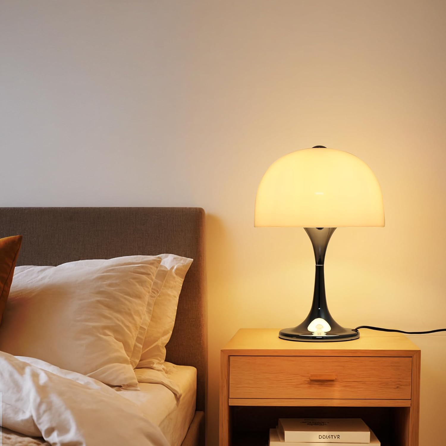 Una lámpara de mesa con base cromada y pantalla blanca en forma de cúpula, sobre una mesita de noche junto a una cama.
