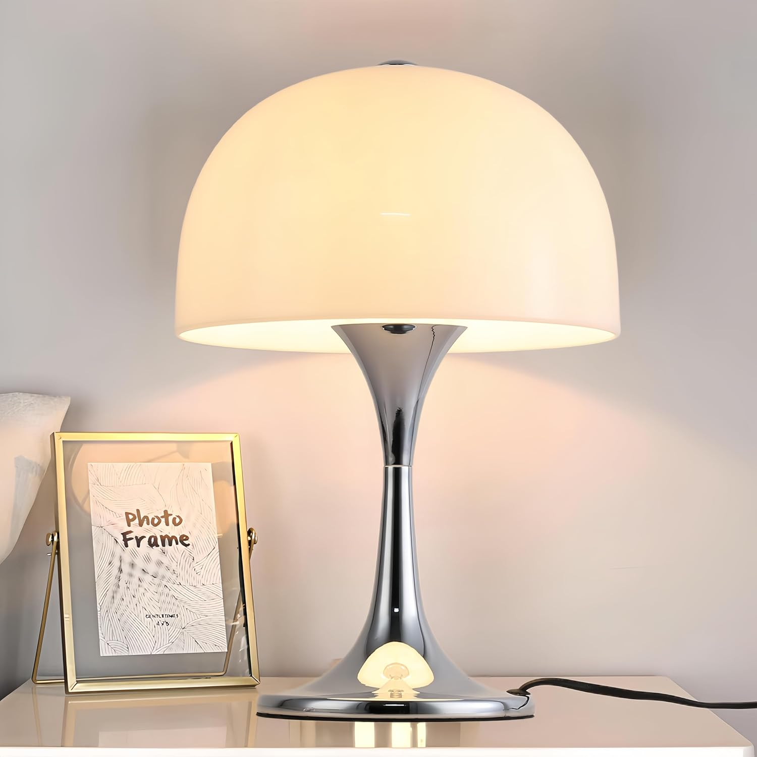 Una elegante lámpara de mesa con base cromada y pantalla blanca en forma de cúpula, sobre una mesita de noche.