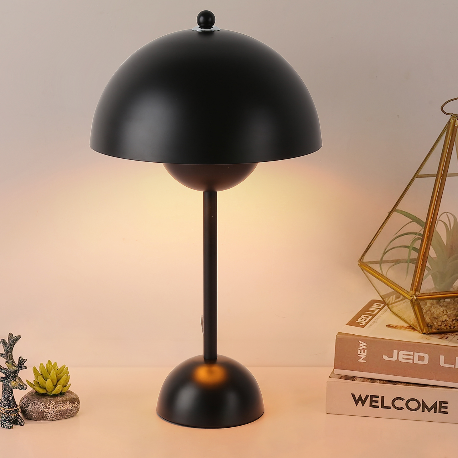 Lámpara de mesa negra con pantalla de cúpula redondeada. Se encuentra sobre una superficie blanca con libros y plantas.