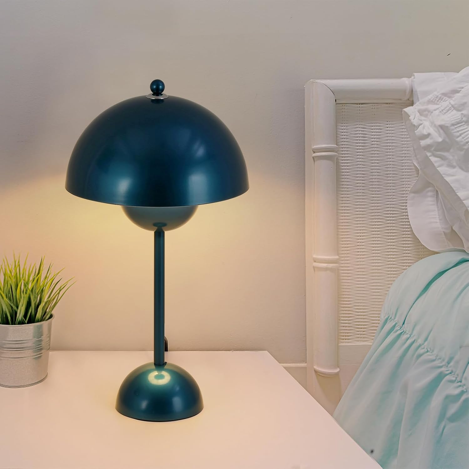 Lámpara de mesa azul encendida sobre mesa blanca, con planta y cama al fondo.