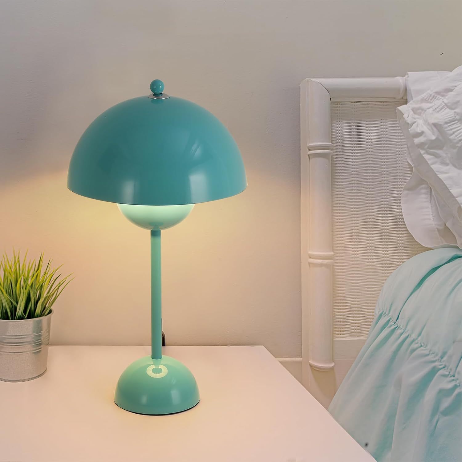 Lámpara de mesa azul encendida sobre mesa blanca, con planta y cama al fondo.