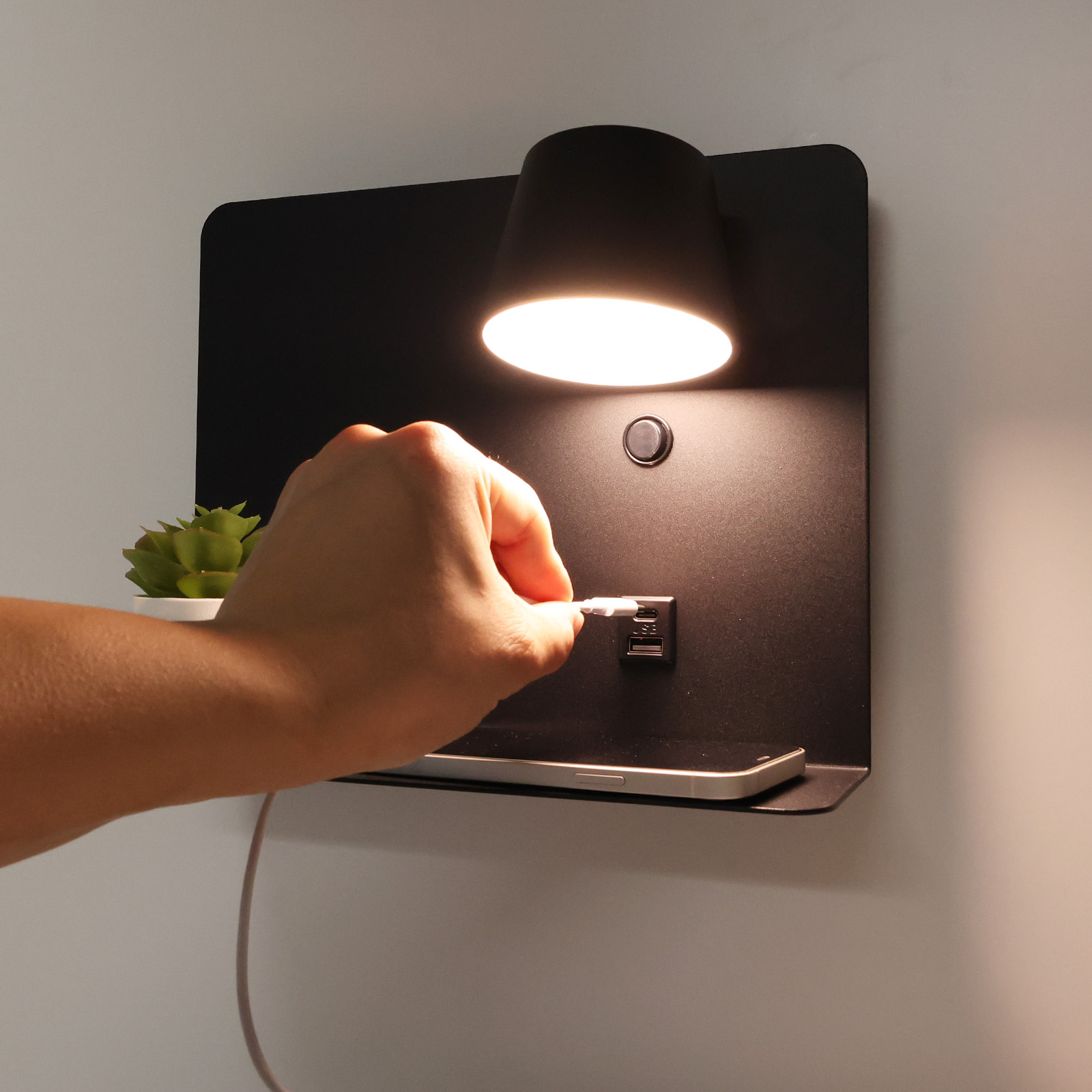 Una mano enchufando un cable USB-C en una lámpara negra montada en la pared con un estante, un teléfono y una planta.
