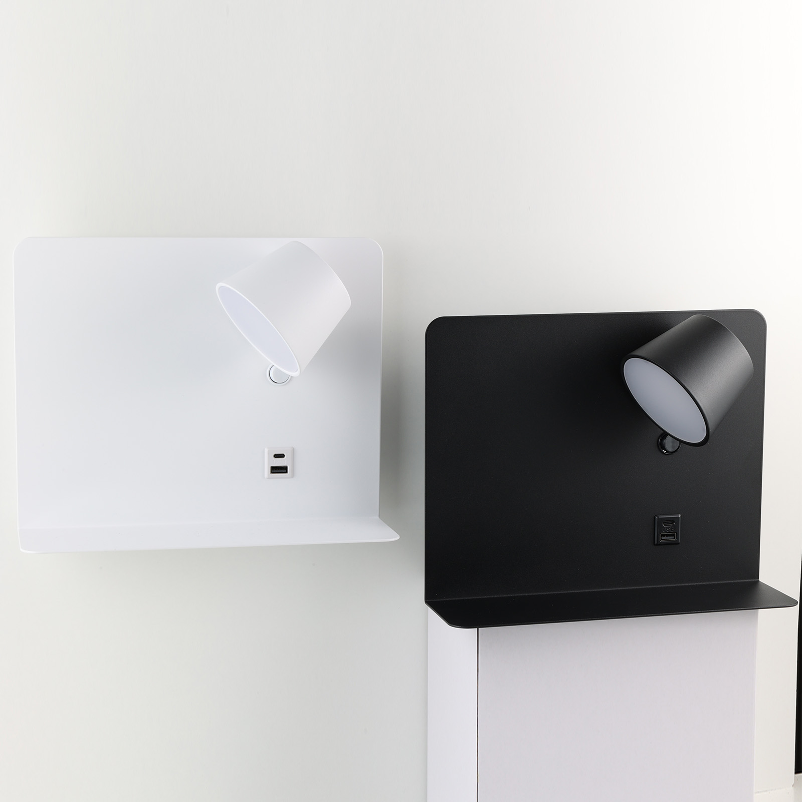 Dos lámparas de pared, una blanca y otra negra, cada una con un foco y puertos USB, montadas en una pared.