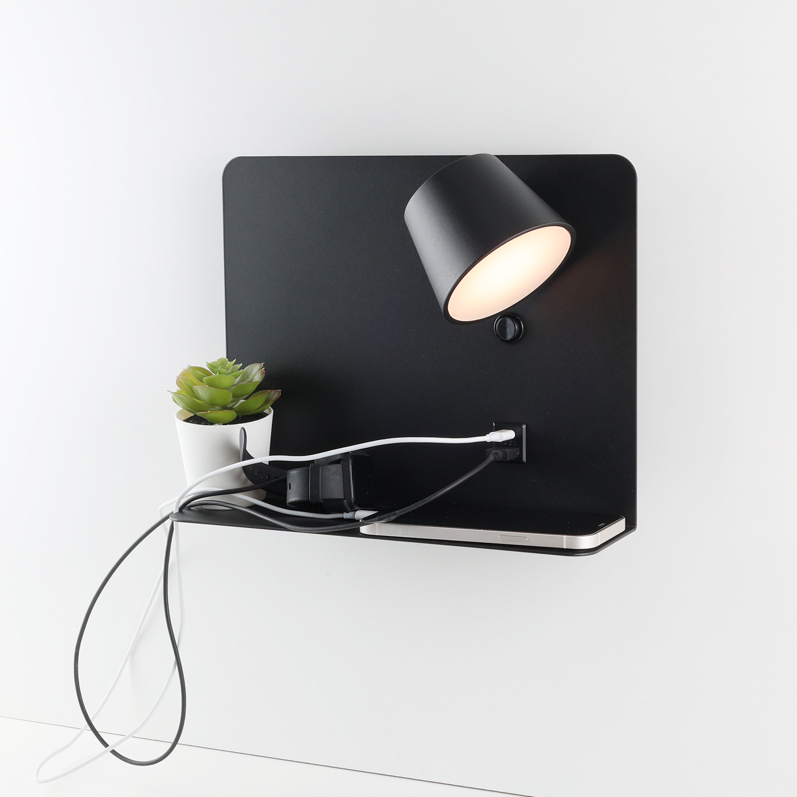 Lámpara de pared negra con un foco, una pequeña planta y cables de carga, montada en una pared blanca.