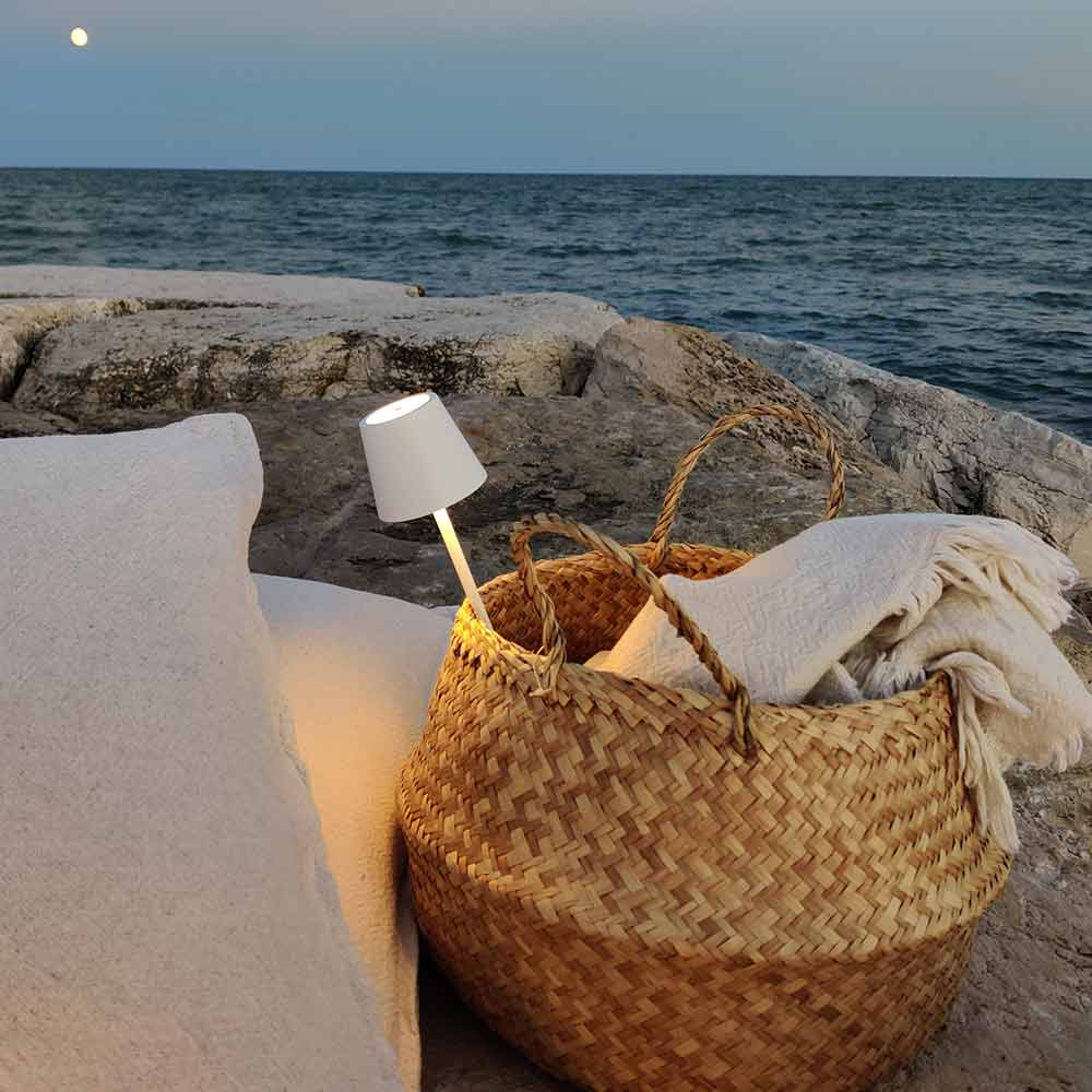Una cesta de mimbre con una manta blanca y una lámpara blanca en una playa rocosa cerca del mar.