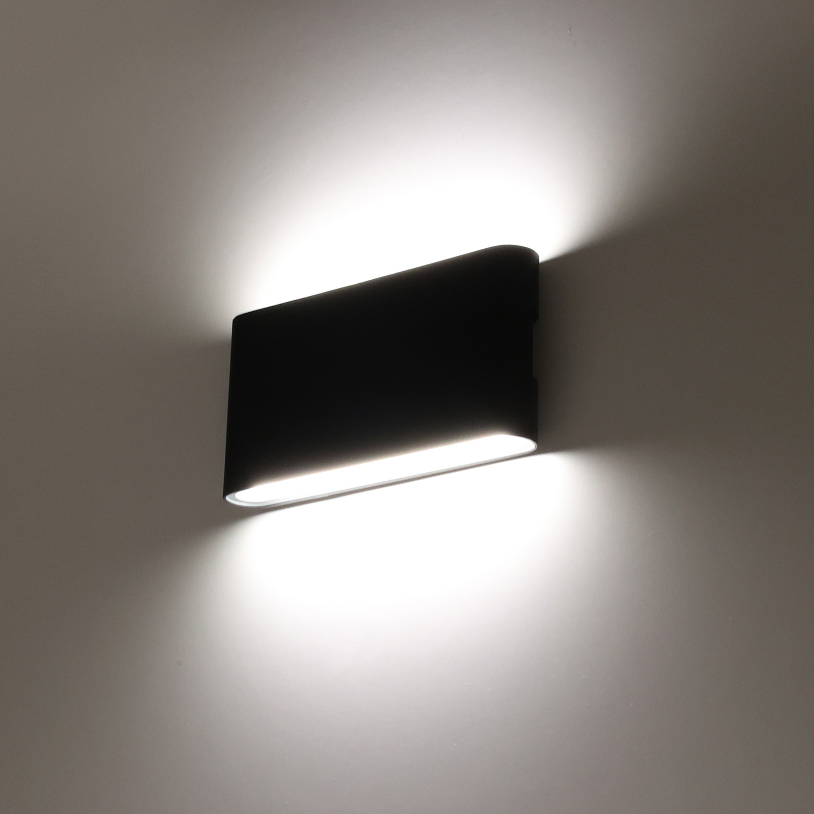 Una lámpara de pared rectangular negra montada en una pared blanca. La luz emana de la lámpara.