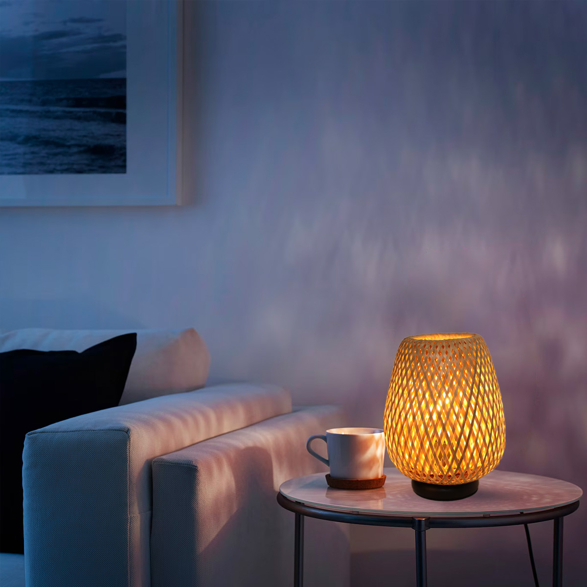Una lámpara de bambú encendida en una mesa, junto a un sofá, en una habitación con un cuadro en la pared.
