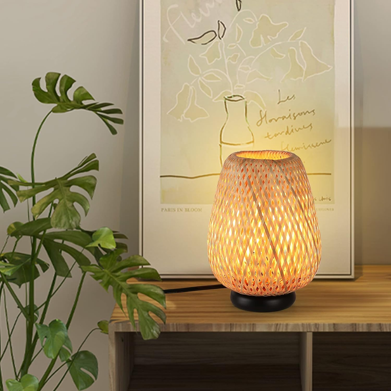 Una lámpara de ratán sobre una mesa de madera, con una pintura y una planta de fondo. La lámpara está encendida.