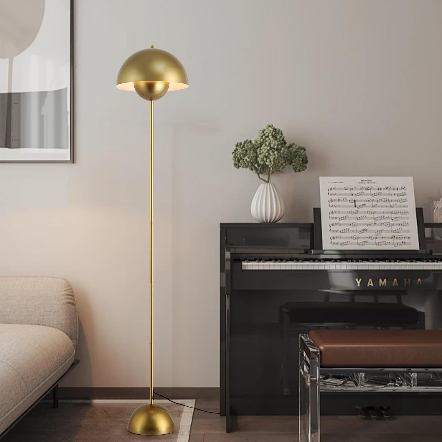 Una lámpara de pie dorada con una pantalla redondeada, iluminando una habitación. Piano, arte, jarrón y sofá en el fondo.