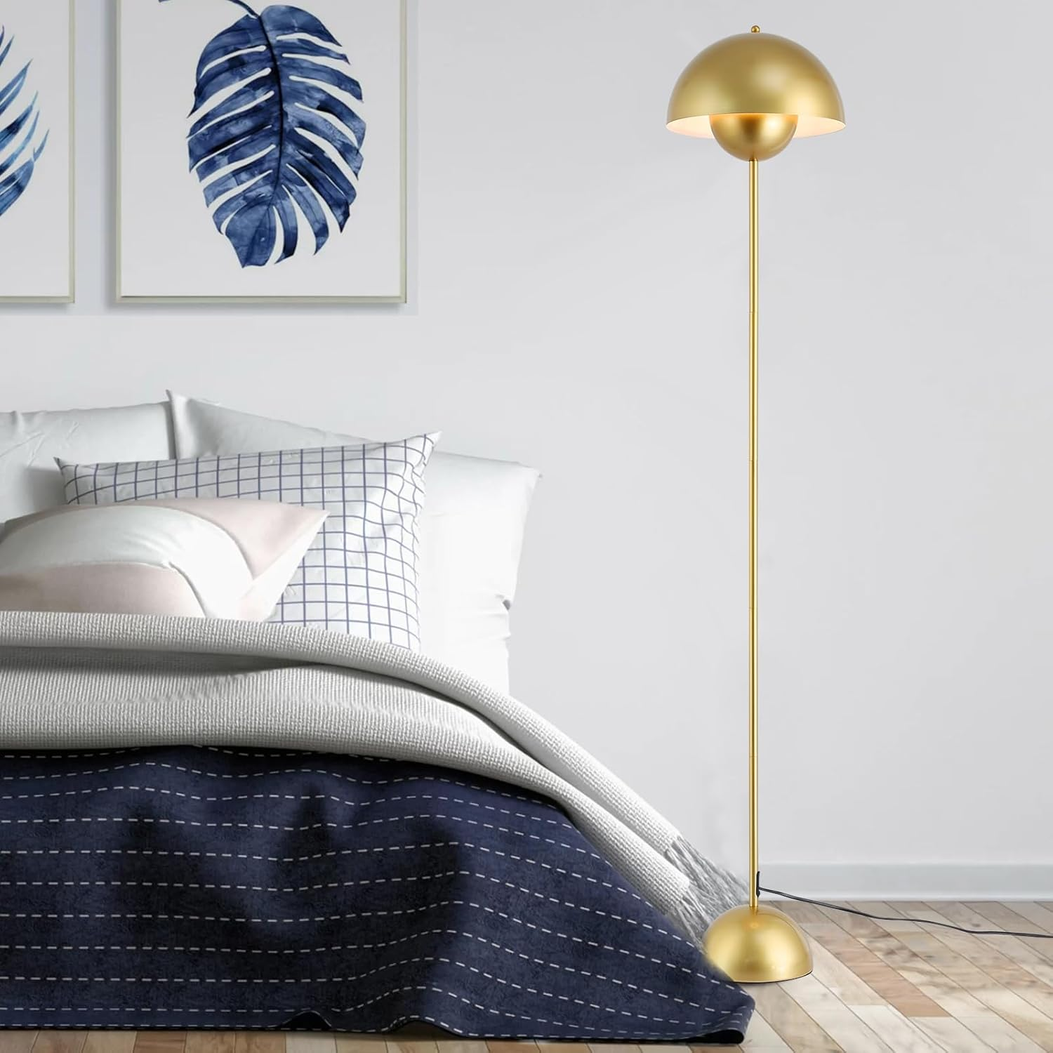 Lámpara de pie dorada con pantalla abovedada, colocada cerca de una cama con ropa de cama azul y un cuadro enmarcado.