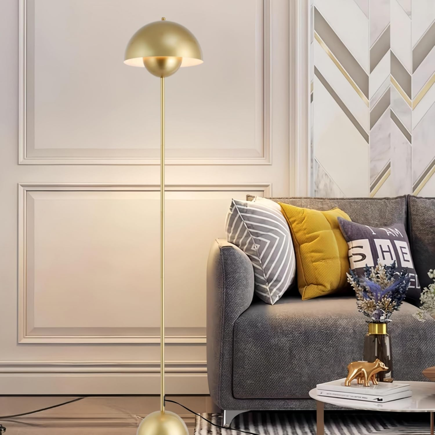 Una lámpara de pie dorada con una pantalla redondeada, iluminando una habitación con un sofá gris, almohadas y una mesa auxiliar.
