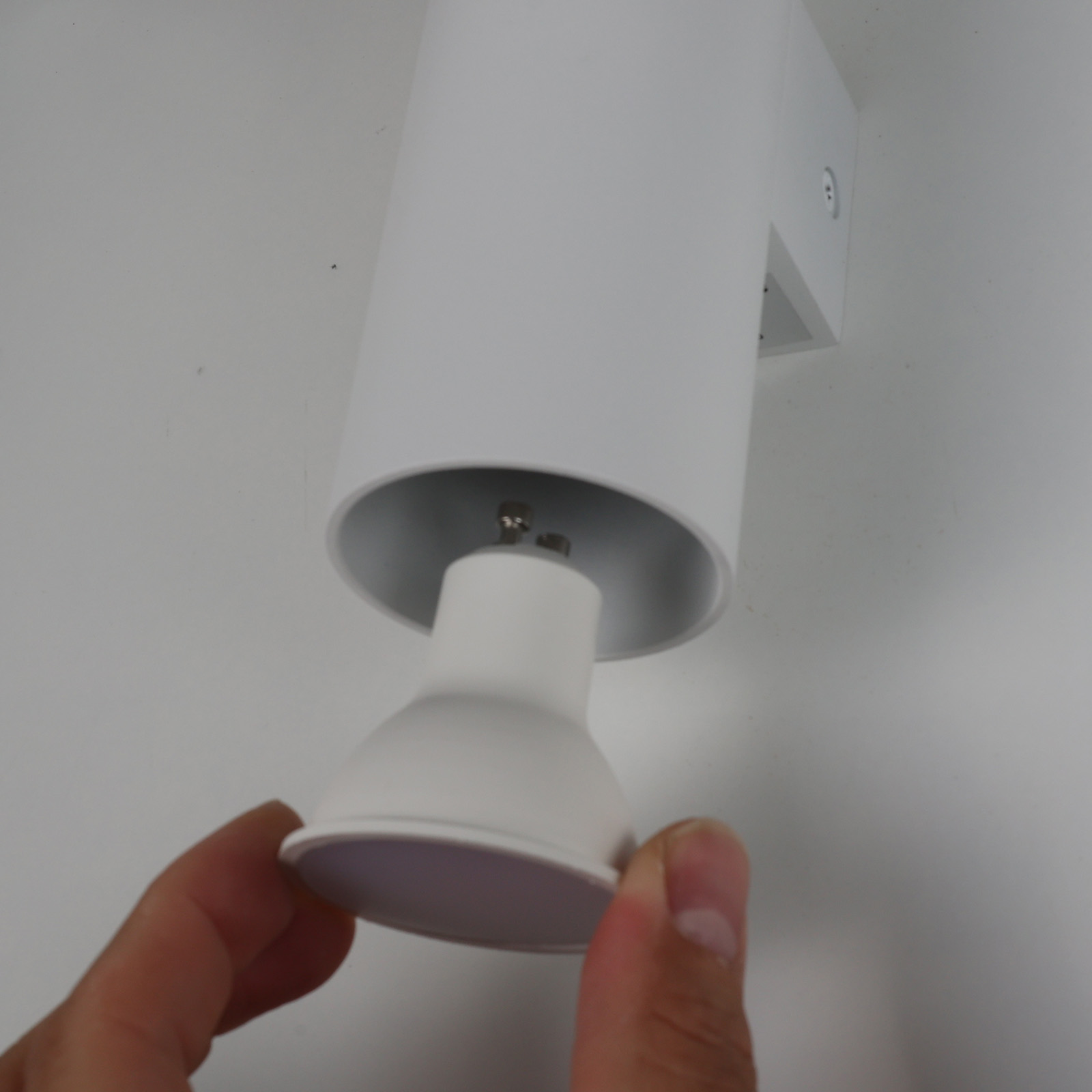 Una mano instala una bombilla GU10 blanca en una lámpara de pared cilíndrica blanca. Montada en una pared blanca.