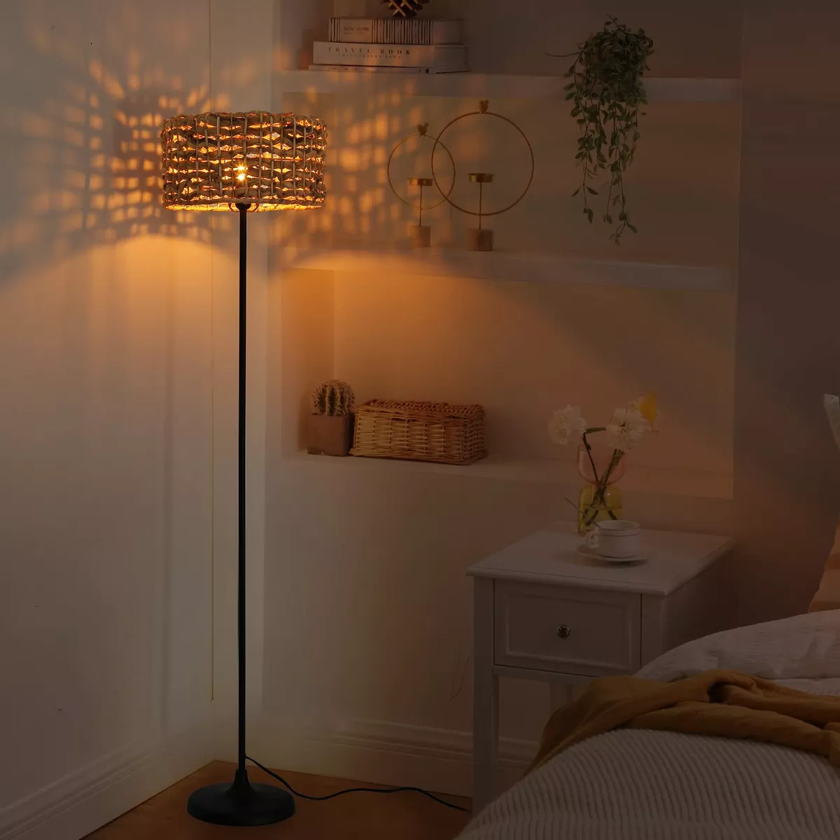 Una lámpara tejida se encuentra en un soporte negro en una habitación. La habitación tiene una cama, una mesita de noche y un cuadro.