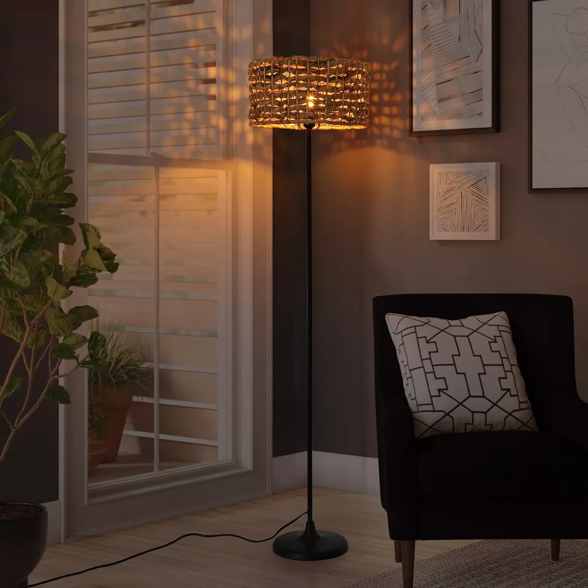 Lámpara tejida sobre un soporte negro. La lámpara ilumina una habitación con ventana, silla y arte enmarcado.