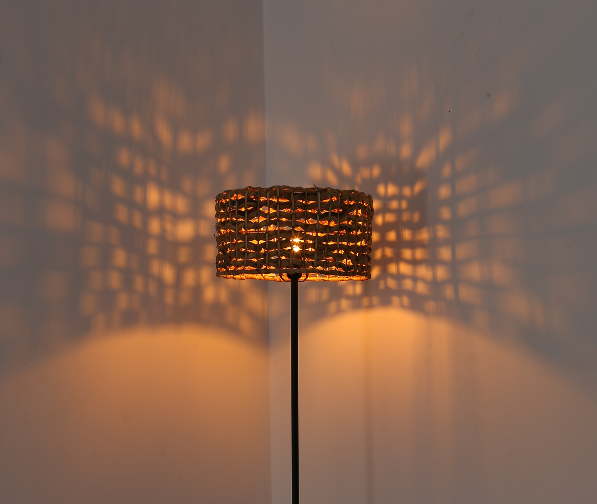 Lámpara de pie con pantalla tejida, proyectando un patrón en la pared. Luz cálida, creando efectos de sombra.