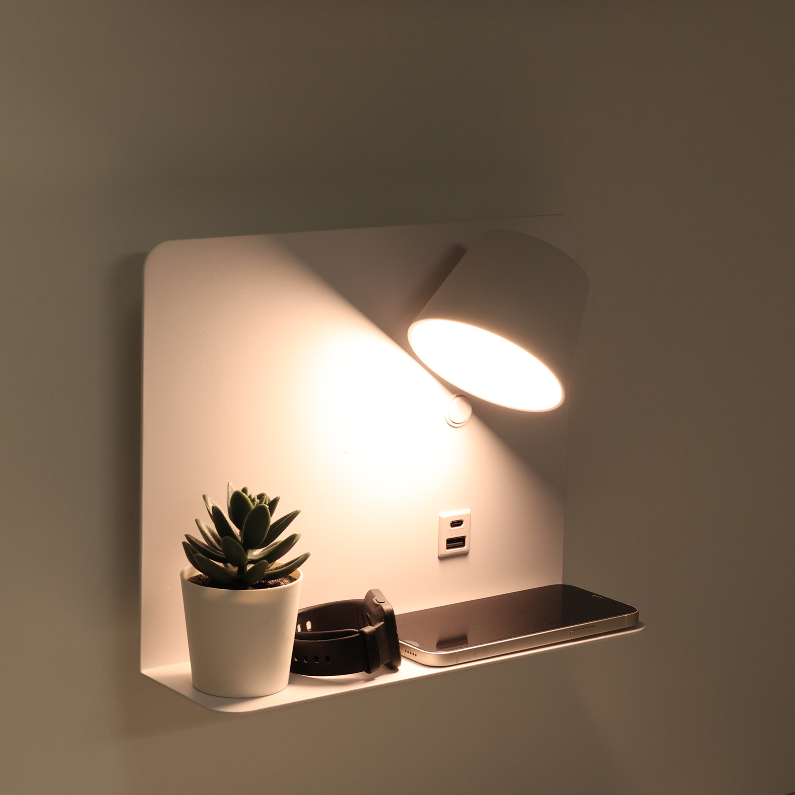 Una lámpara blanca montada en la pared con un estante, una planta, un reloj y un teléfono. La lámpara está encendida, iluminando los objetos.