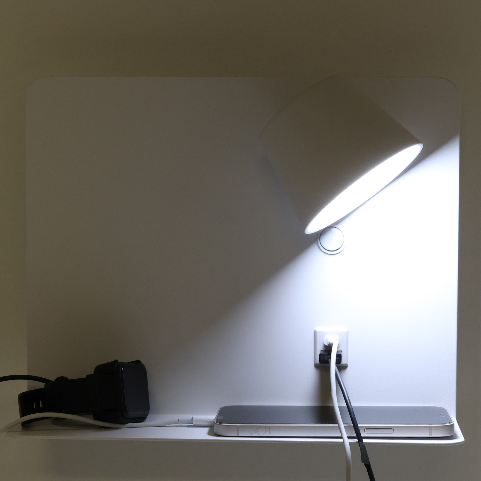 Una lámpara de pared blanca con enchufe de carga y teléfono. La lámpara está encendida.