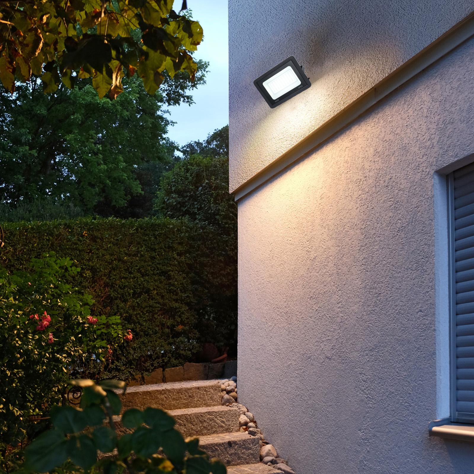 Un foco negro en una pared blanca, junto a una escalera y vegetación. La luz está encendida.