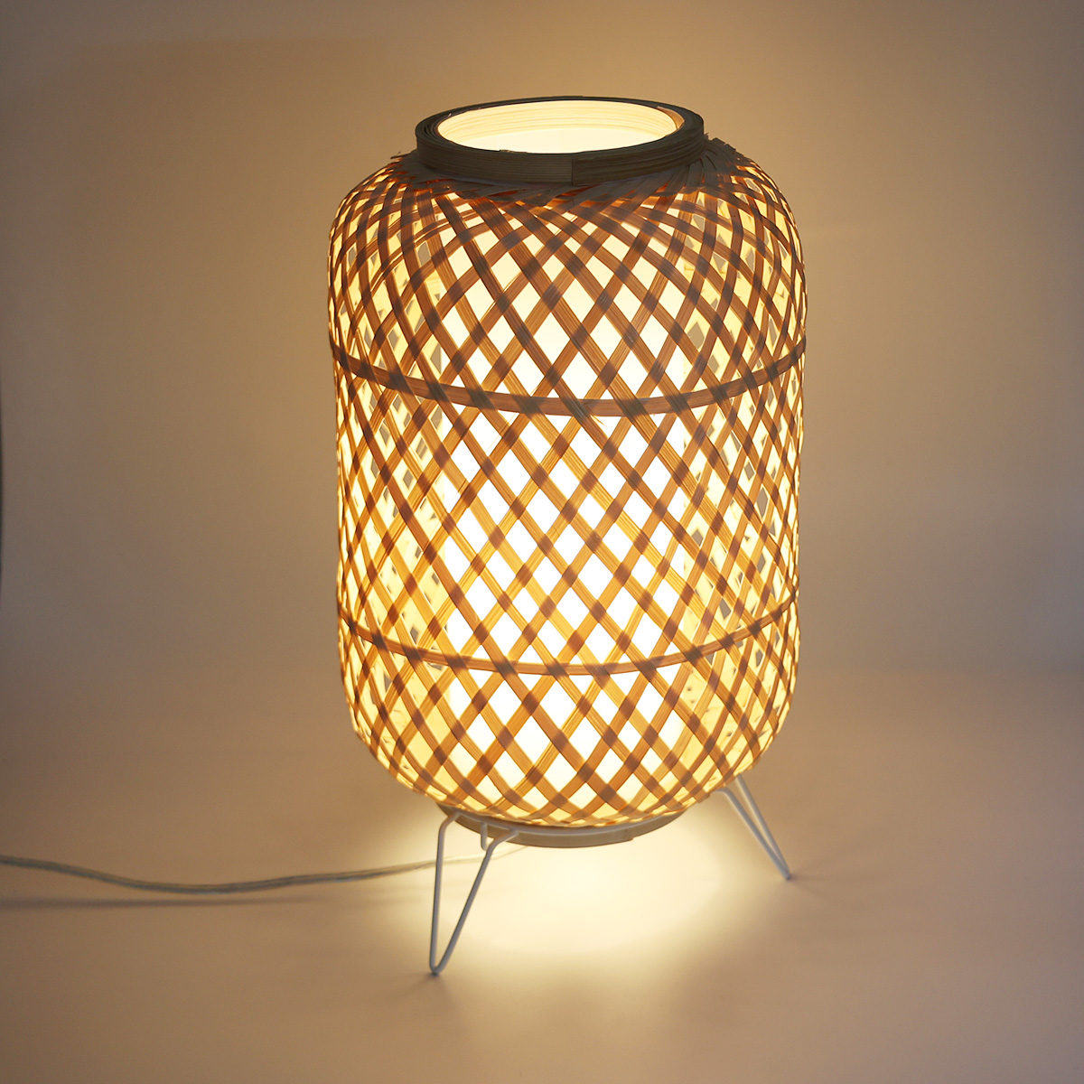 Lámpara cilíndrica de bambú tejido, iluminada y proyectando luz cálida. Tiene un soporte blanco y un cable de alimentación blanco.