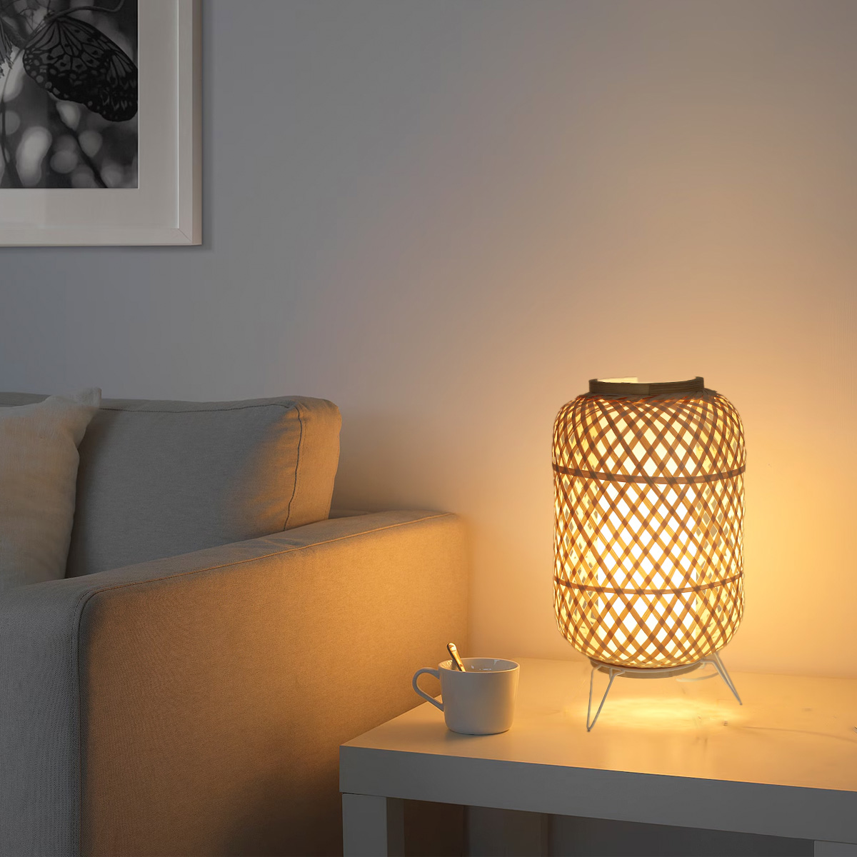Una lámpara de bambú tejido cilíndrica iluminada está sobre una mesa. Un sofá, una taza y un marco de fotos están en el fondo.