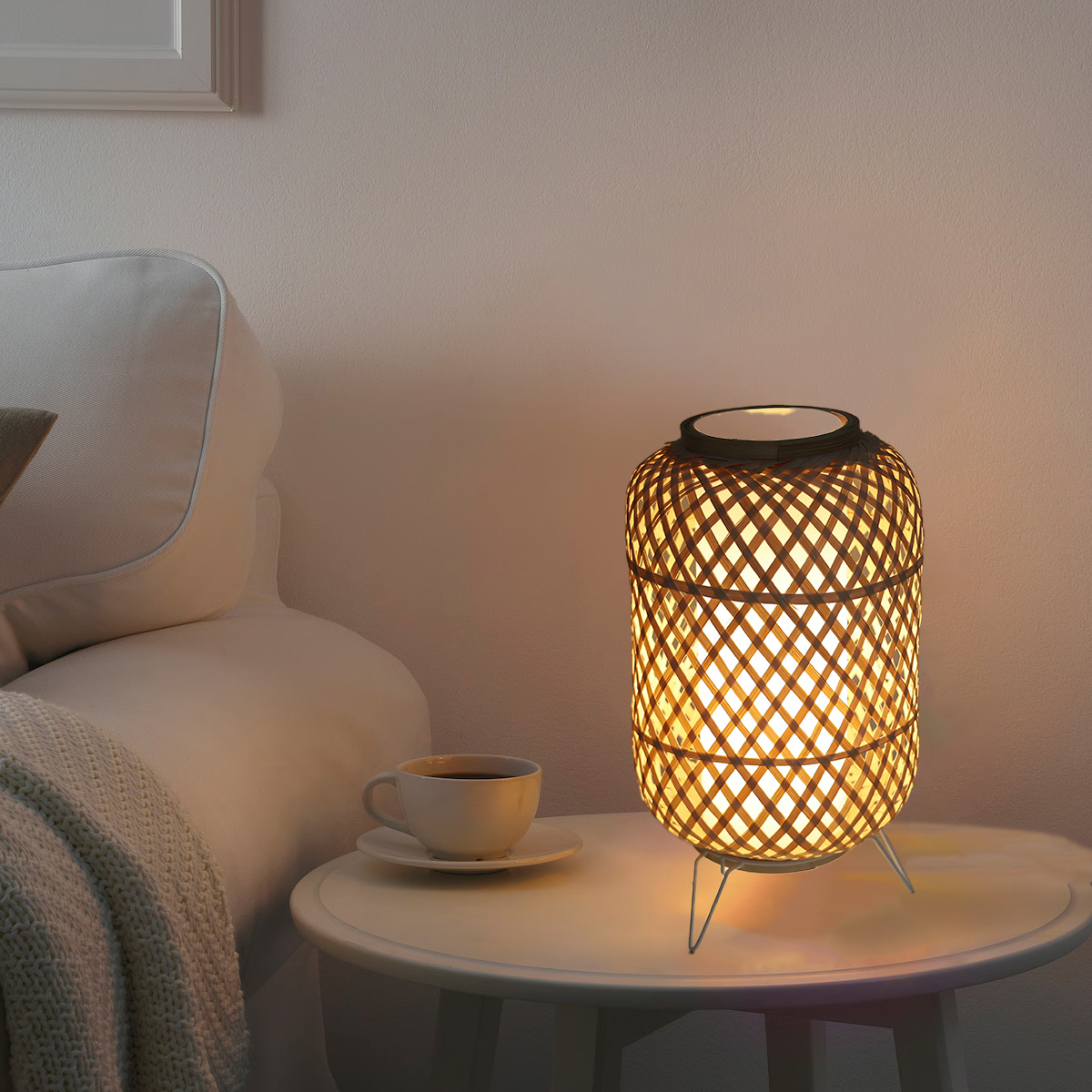 Lámpara de bambú sobre una mesa blanca con taza y platillo. La lámpara está encendida, y una cama al fondo.