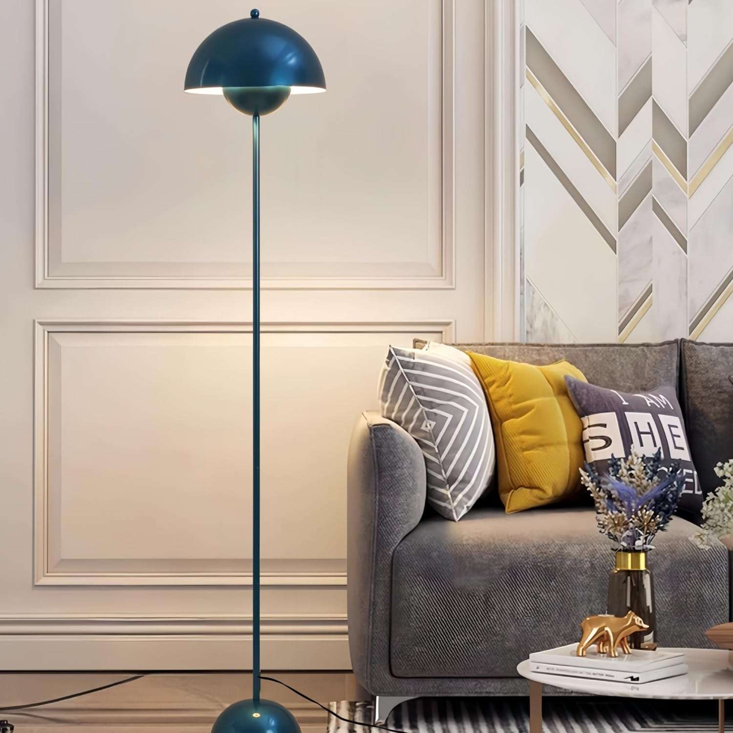 Una lámpara de pie turquesa con pantalla de cúpula junto a un sofá gris con almohadas. El fondo muestra una pared.