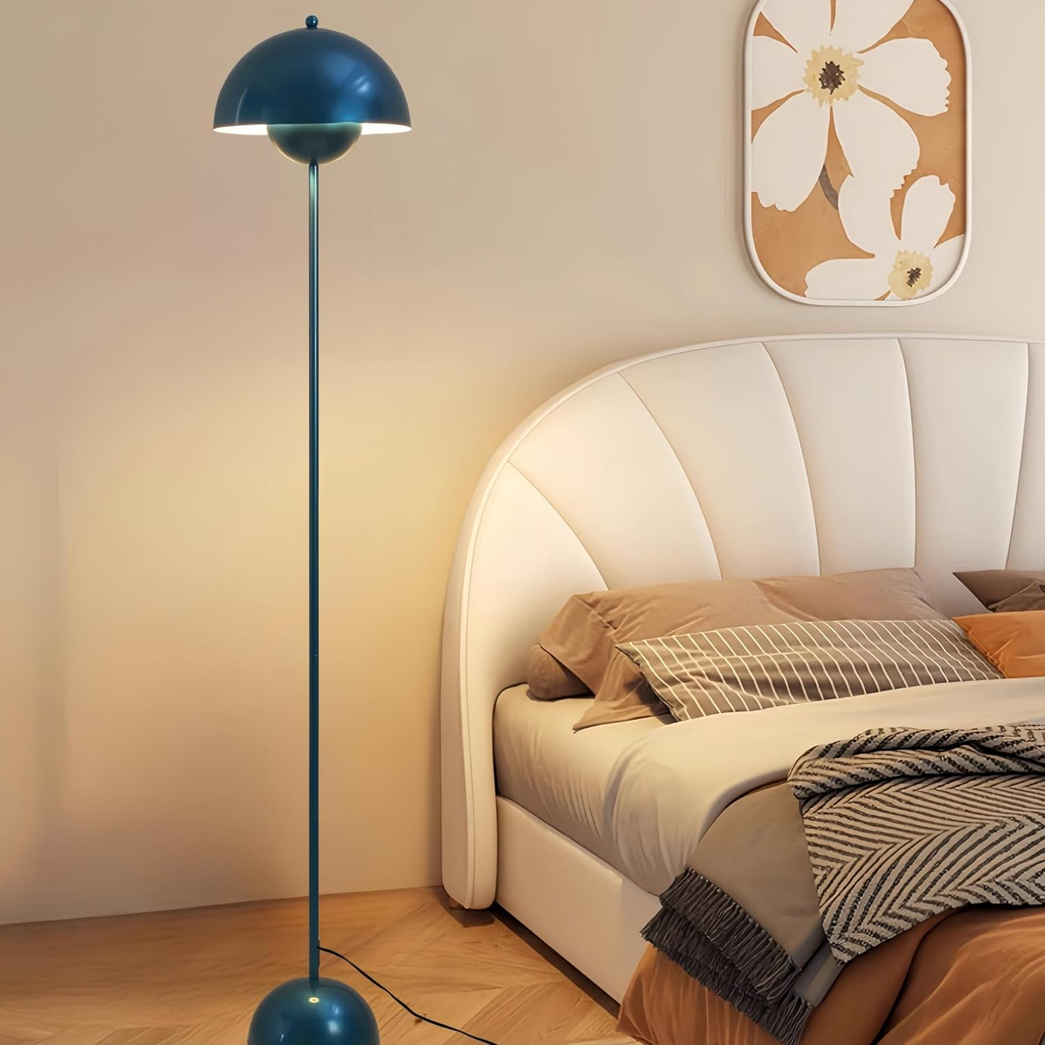 Una lámpara de pie turquesa con pantalla de cúpula en un dormitorio. Una cama con almohadas a la derecha. Un cuadro sobre la cama.