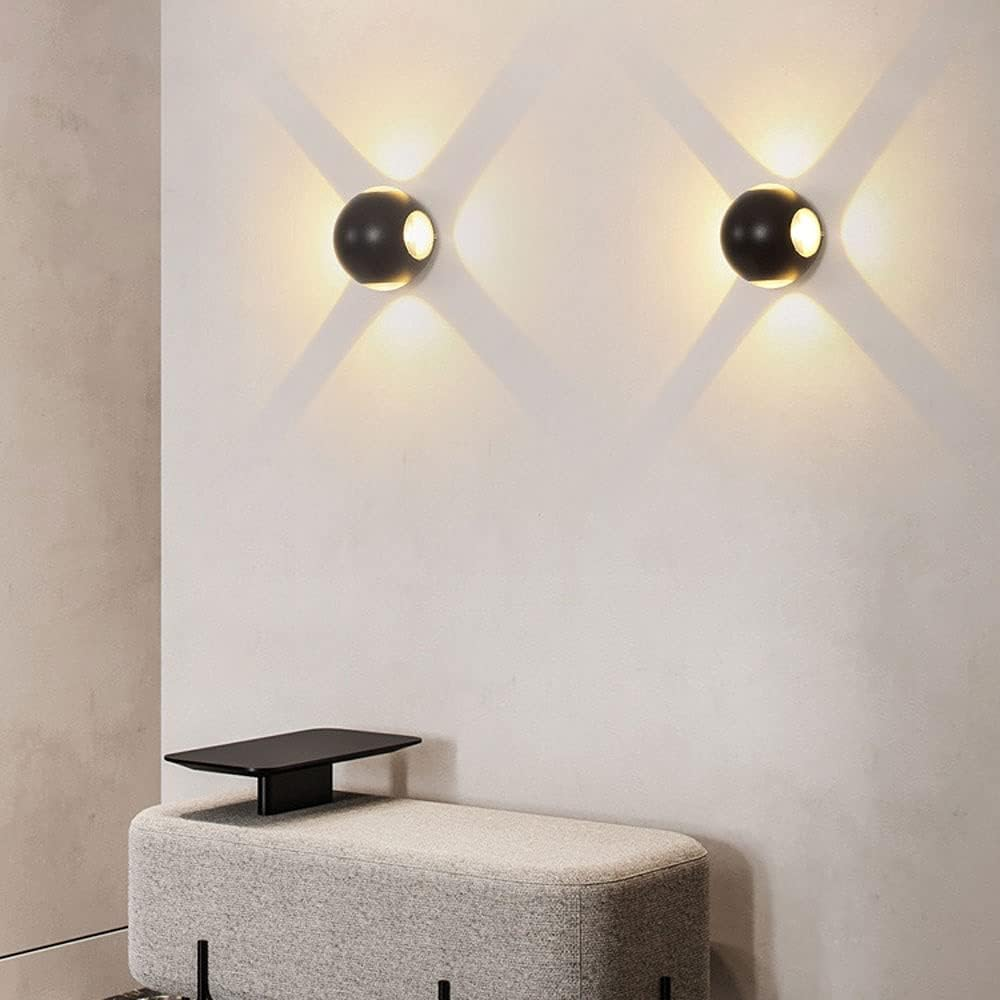 Dos lámparas de pared negras que emiten luz, montadas en una pared blanca sobre un banco gris. Sombras de luz presentes.
