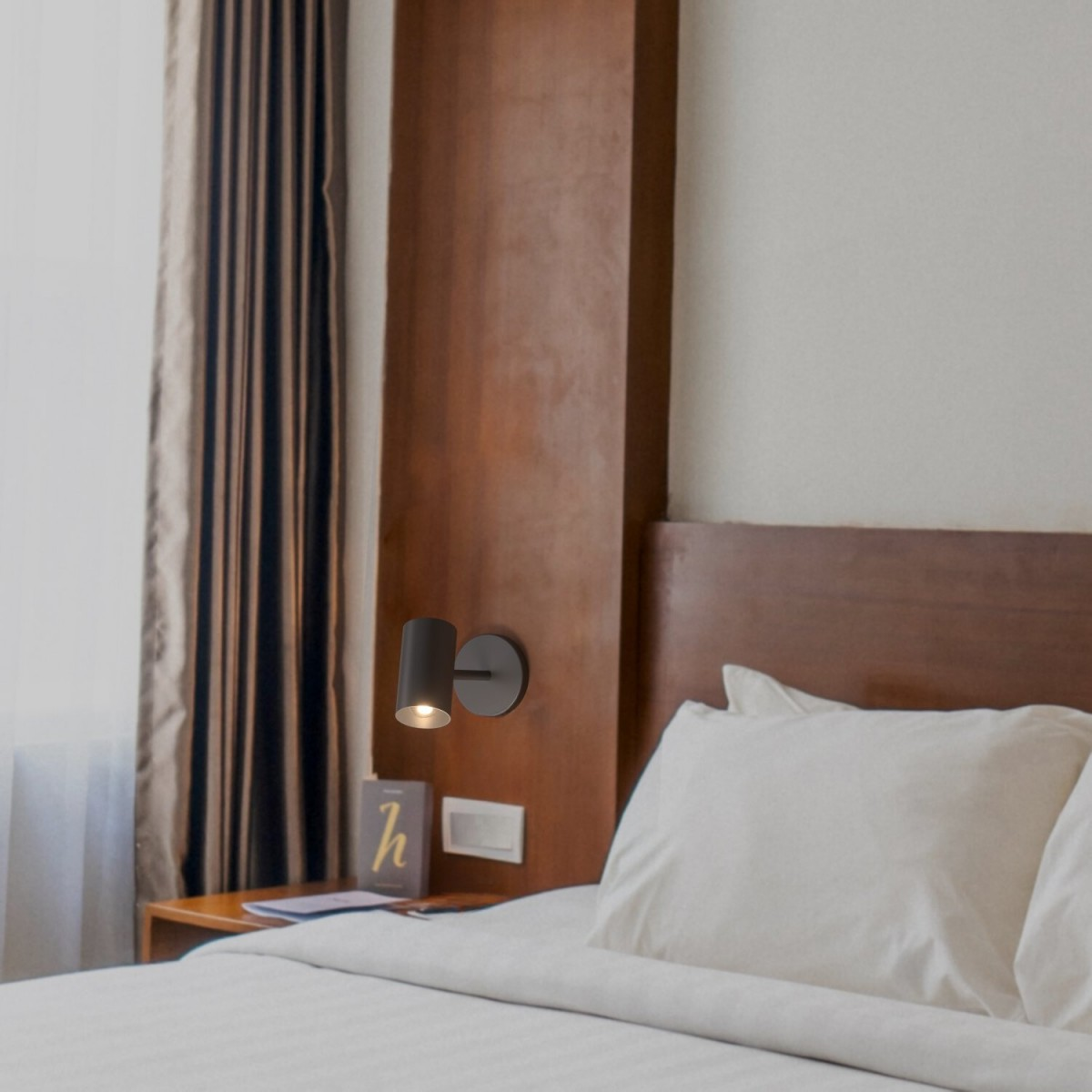 Lámpara de pared negra junto a una cama, iluminando un área pequeña con luz cálida.
