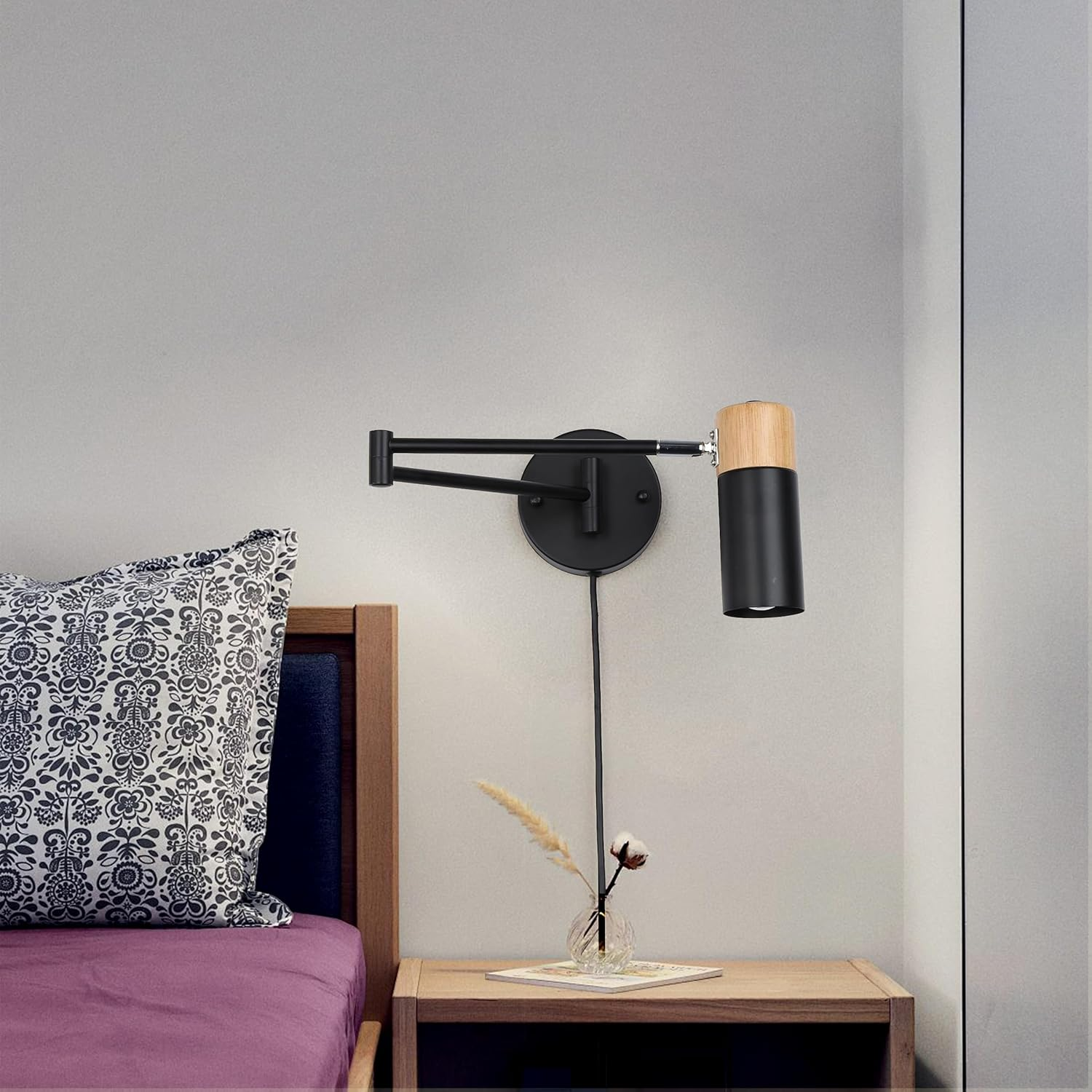 Una lámpara de pared negra y de madera está montada en una pared blanca junto a una cama. Se ve una mesa pequeña con decoración.
