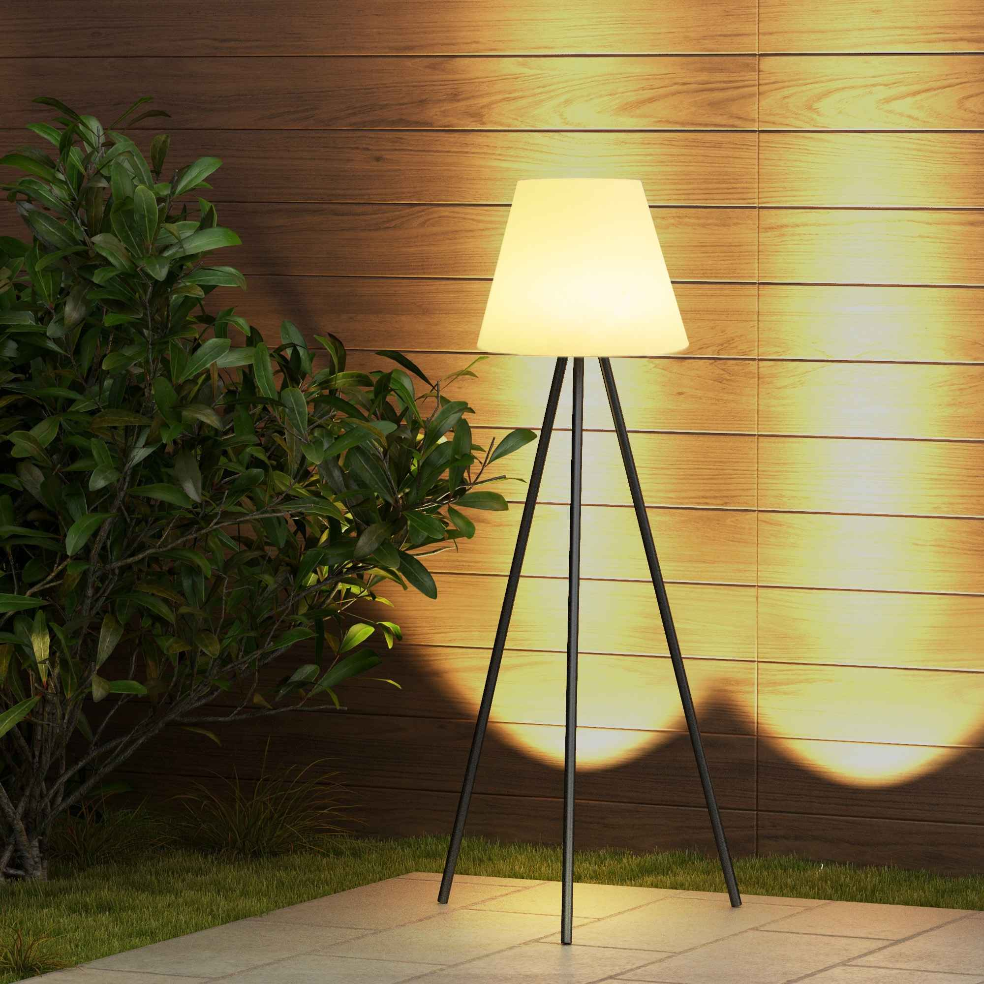 Stehlampe mit weißem, konischem Schirm und schwarzen Beinen, vor einer Holzwand und einer grünen Pflanze.