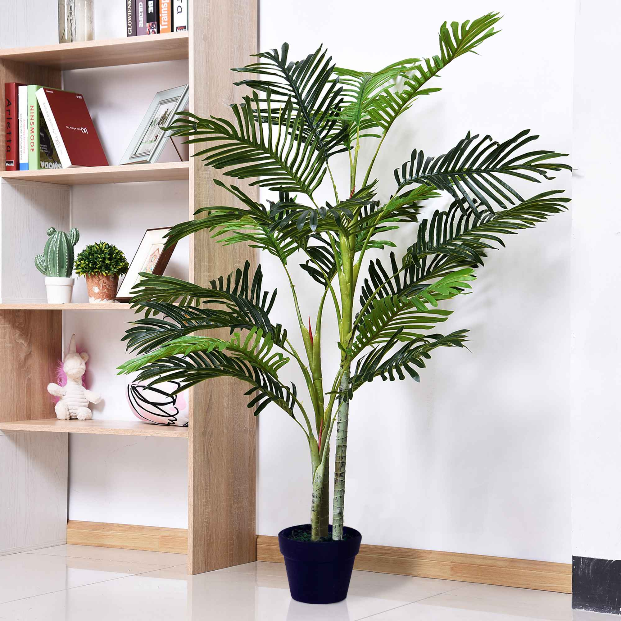 Eine hohe künstliche Palme in einem dunklen Topf, mit einem Bücherregal im Hintergrund.
