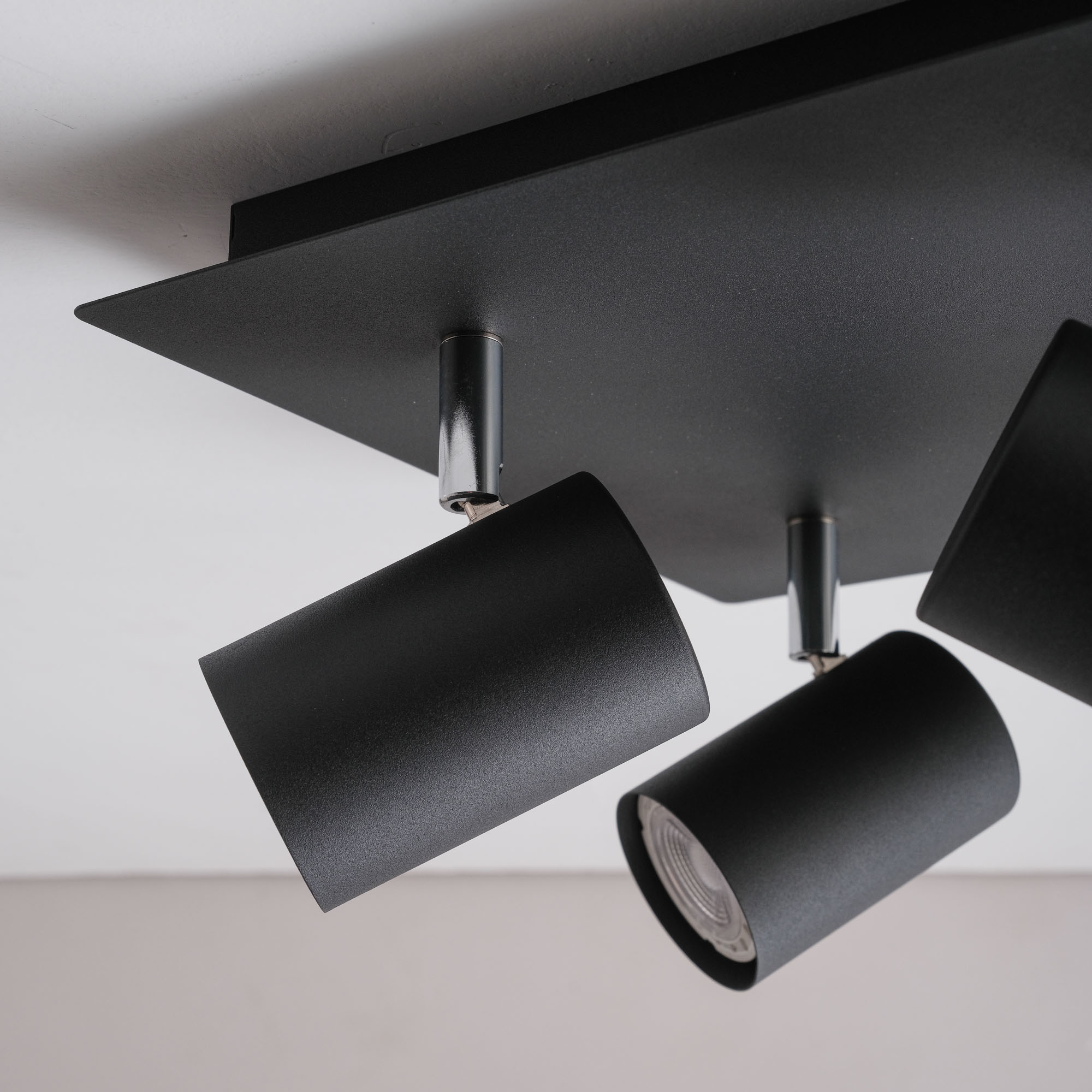Schwarzer Deckenstrahler mit drei zylindrischen Lampen. Weißer und hellgrauer Hintergrund.