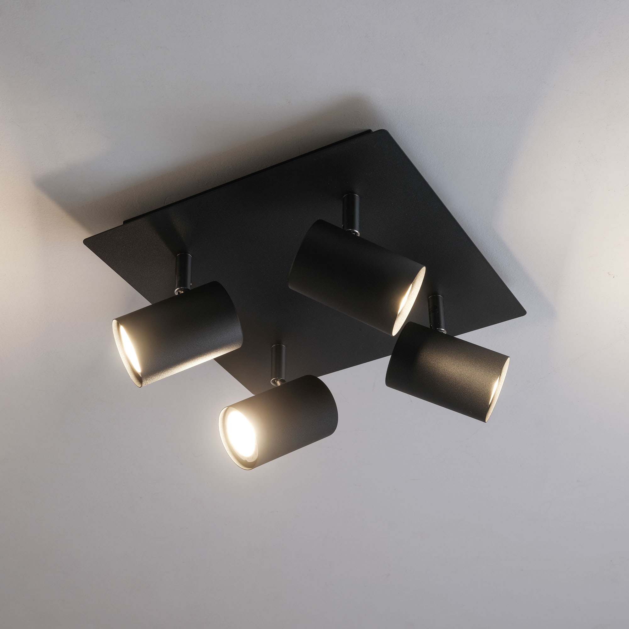 Schwarze Deckenleuchte mit vier Strahlern, vor weißer Decke. Das Licht ist an.