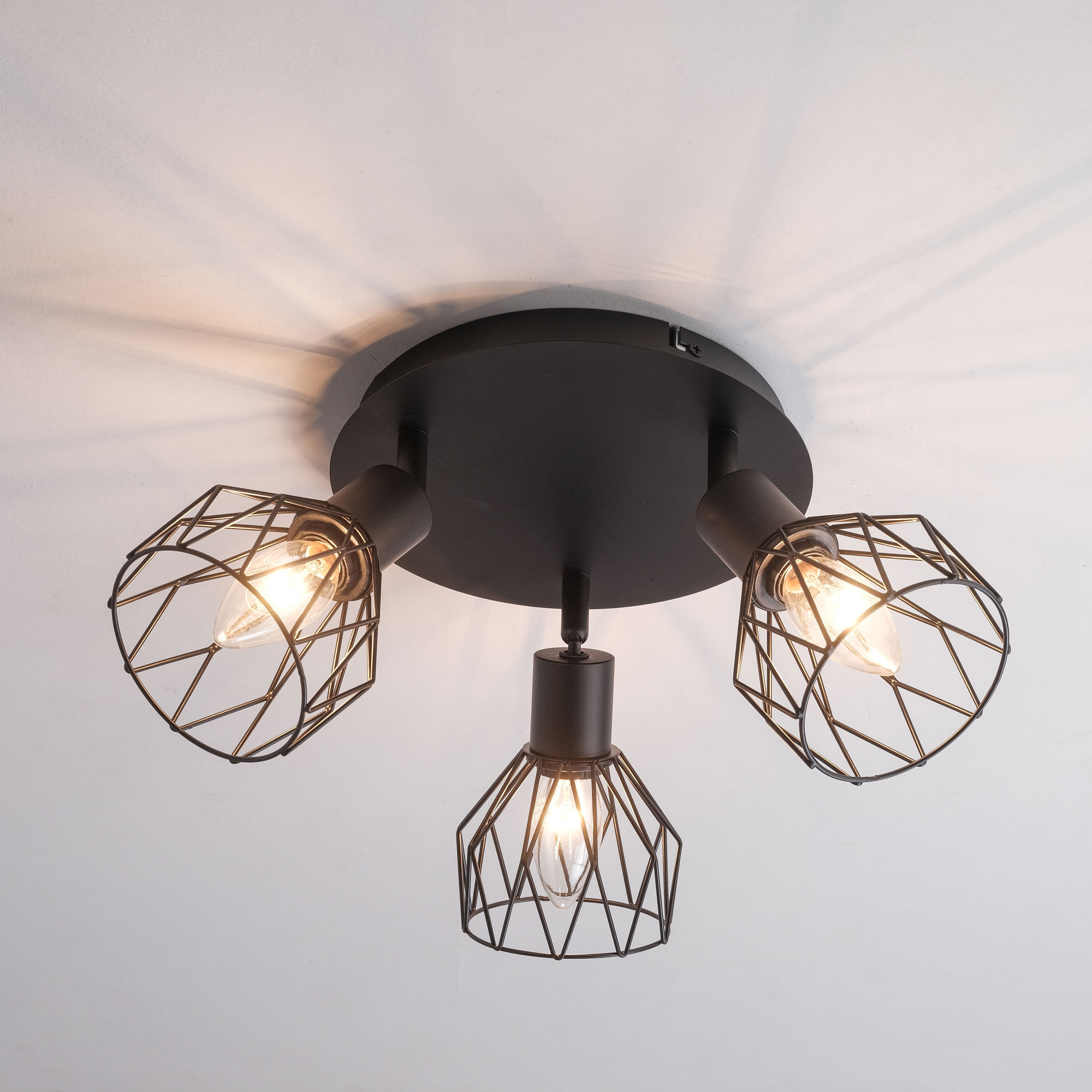 Schwarze Deckenleuchte mit drei Lampenschirmen im Käfigstil. Das Licht ist an einer weißen Decke an.