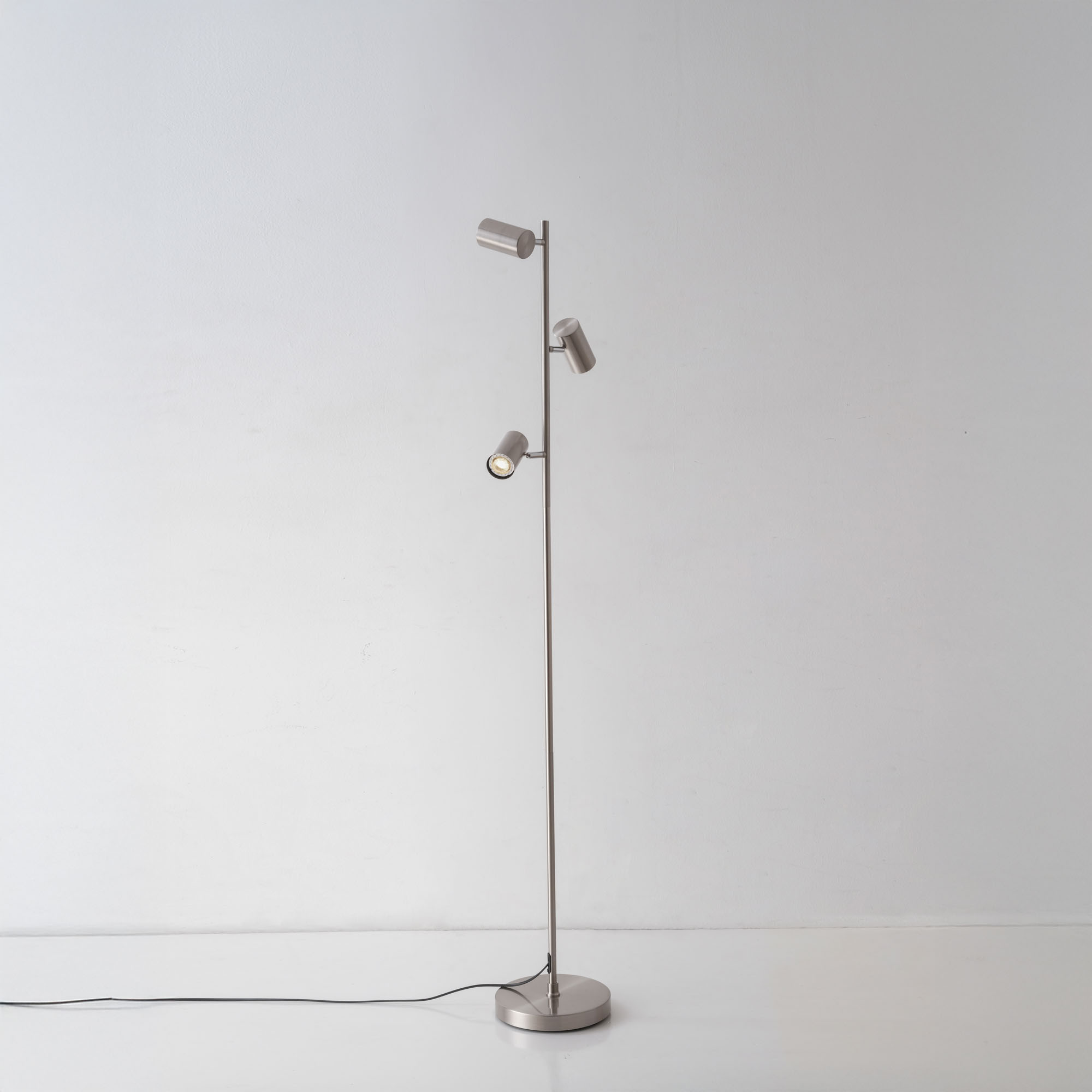 Eine silberne Stehlampe mit drei Strahlern. Das Licht ist vor weißem Hintergrund an.