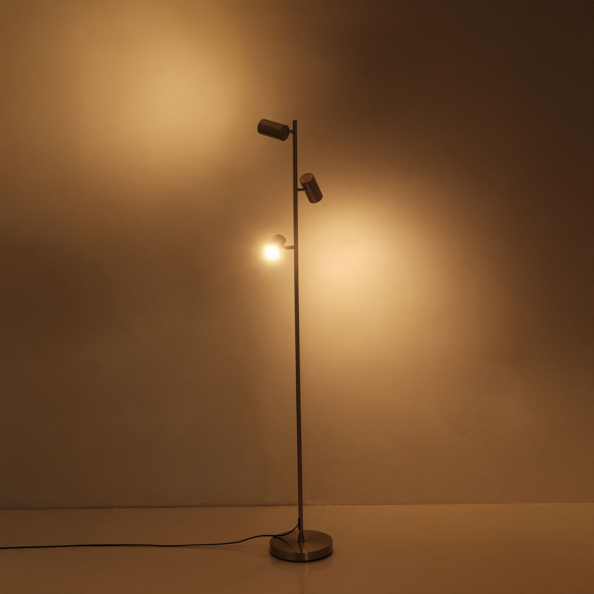 Eine silberne Stehlampe mit drei Strahlern, vor einer warm beleuchteten Wand. Das Licht ist an.