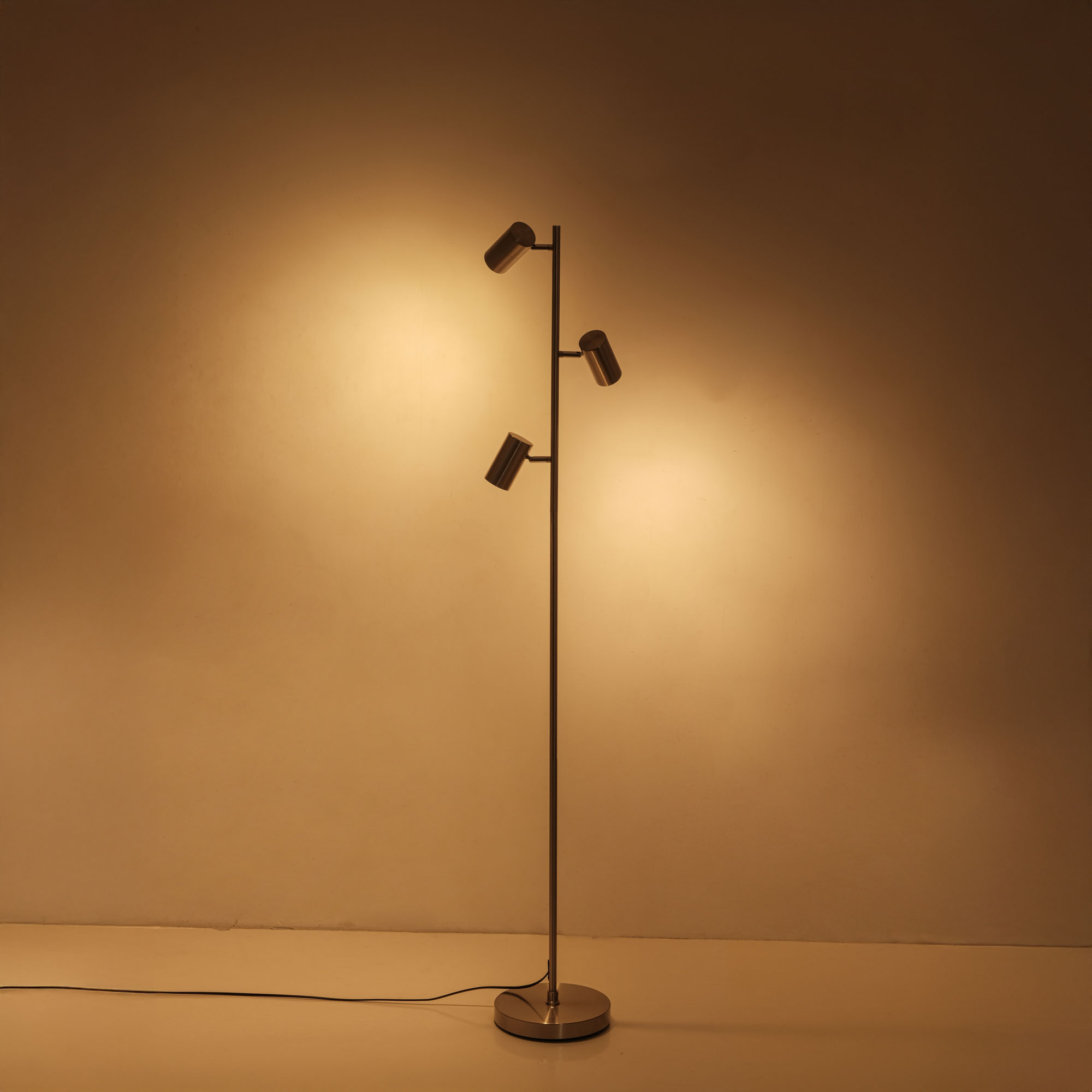 Eine hohe Stehlampe mit drei Strahlern vor einer braunen, beleuchteten Wand.