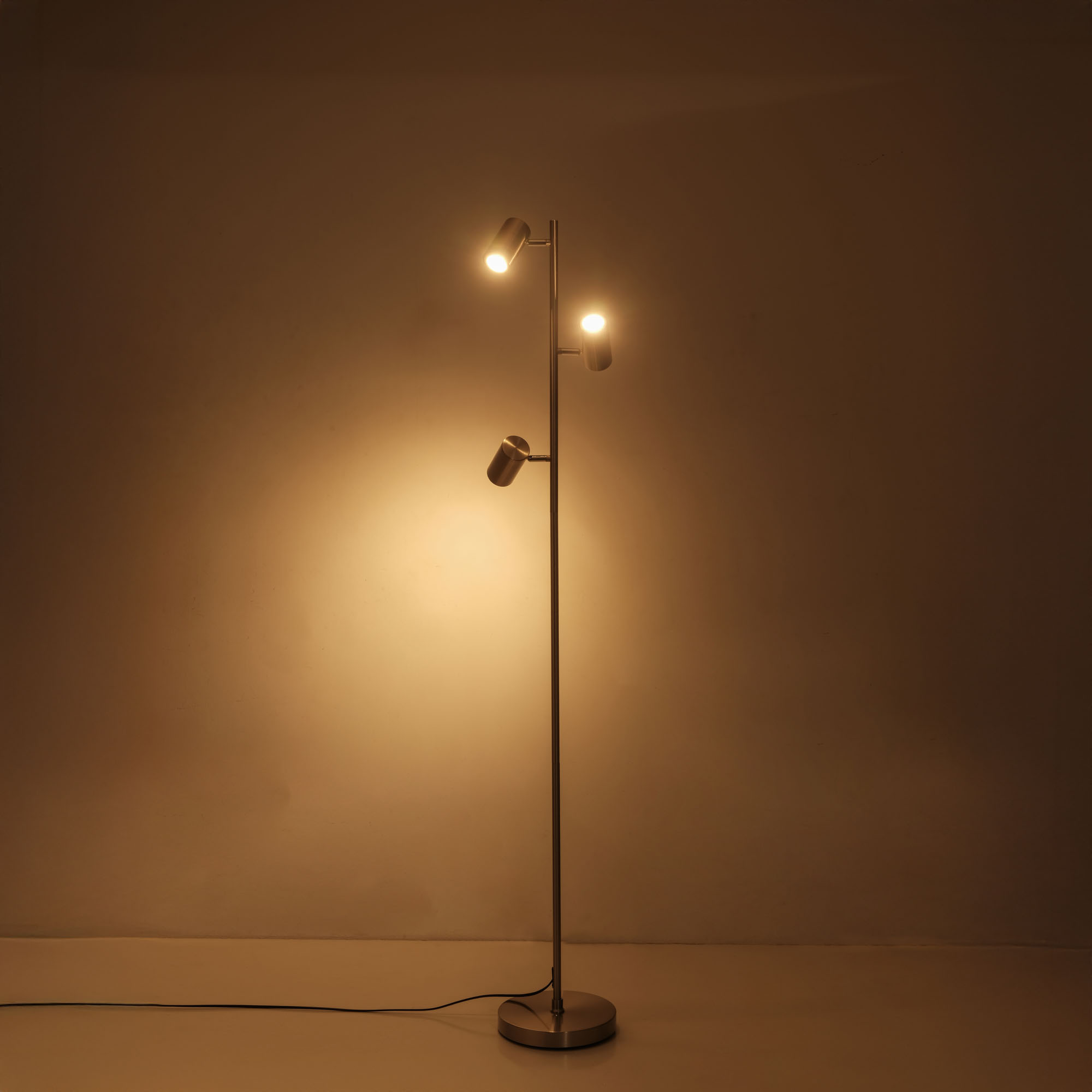 Eine Stehlampe mit drei Strahlern vor einer braunen Wand, die den Bereich beleuchtet.