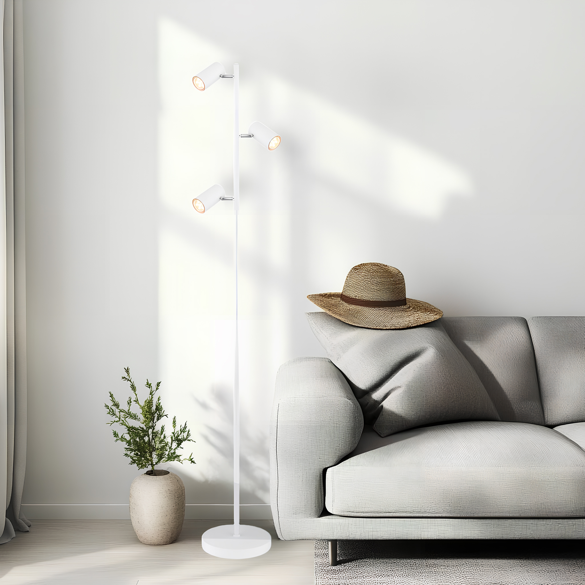 Weiße Stehlampe mit drei Strahlern, ein Hut und ein Sofa im Raum. Das Licht ist an.