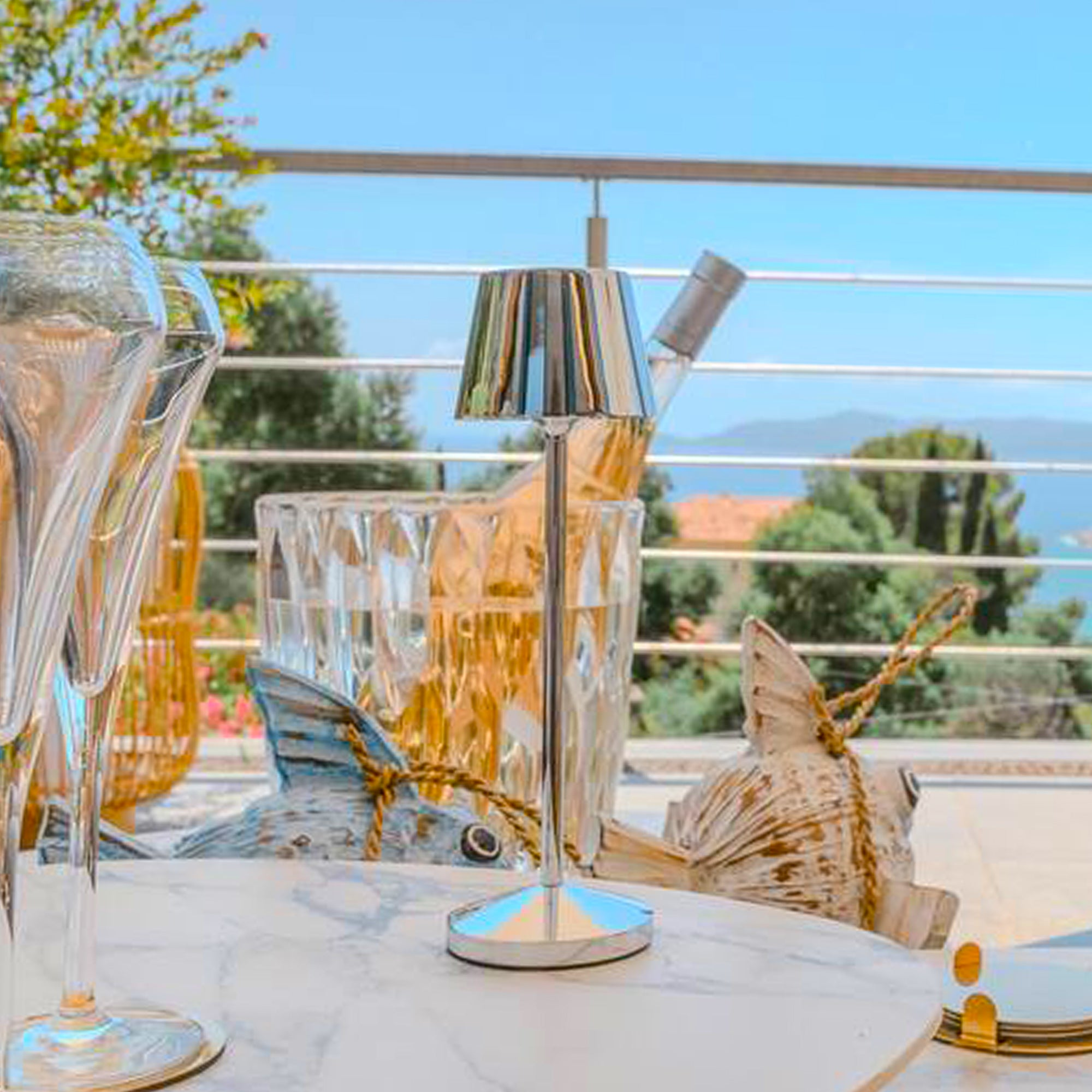 Ein Tisch mit einer Tischlampe, Gläsern und einer Weinflasche mit Blick auf das Meer. Die Lampe ist silber.