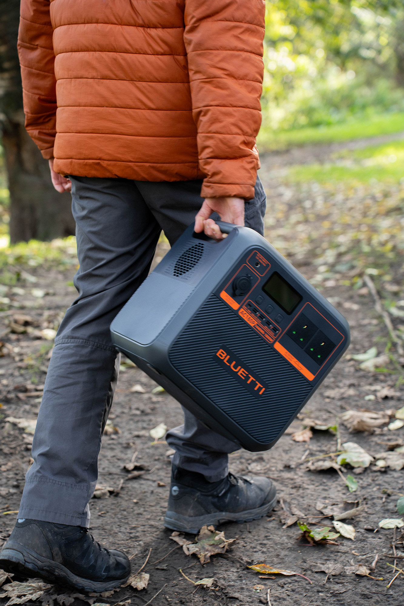 Osoba idąca z przenośną stacją zasilania w parku. Stacja zasilania jest czarna i pomarańczowa.