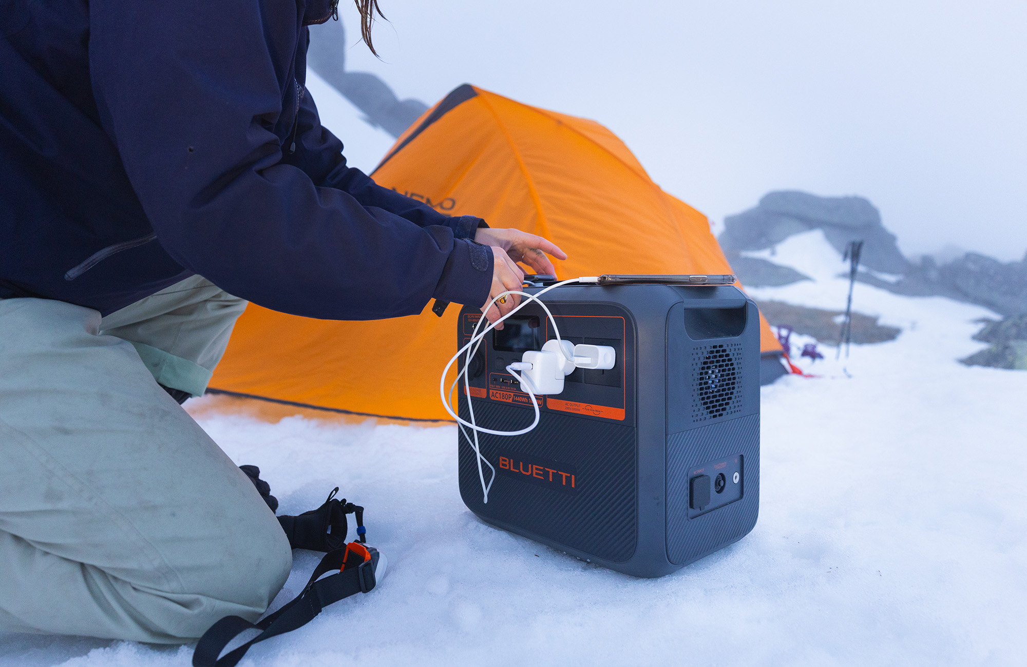 Osoba klęcząca, ładująca urządzenie z czarnej przenośnej stacji zasilania na śniegu. Pomarańczowy namiot w tle.