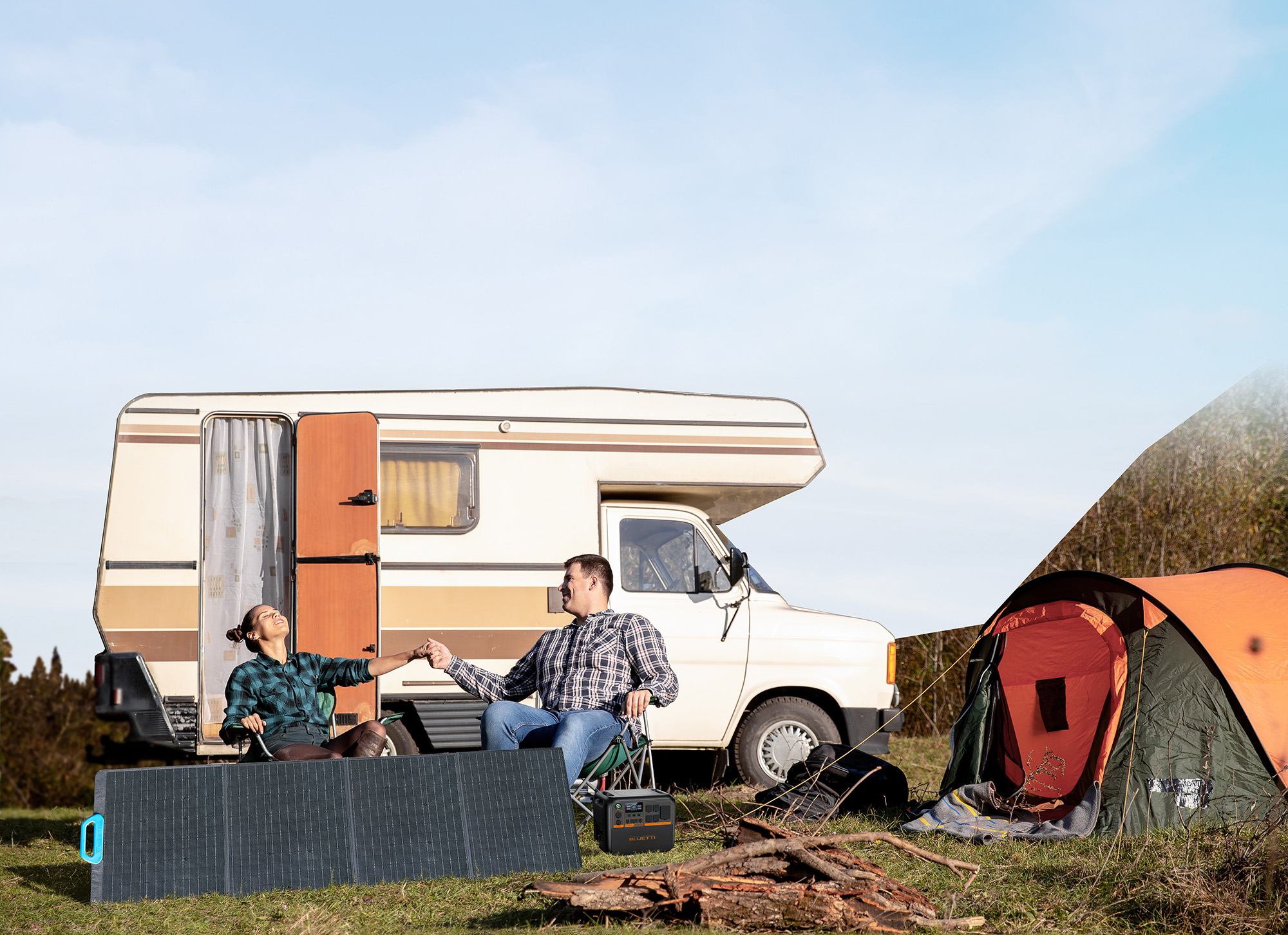 Paar sitzt draußen vor einem Wohnmobil. Ein Campingzelt und ein Solarpanel sind sichtbar.