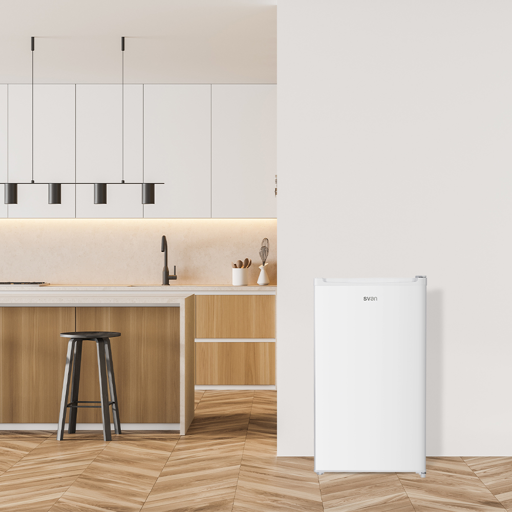 Un refrigerador Svan blanco se encuentra en una cocina moderna con pisos y gabinetes de madera clara, junto a una pared blanca.