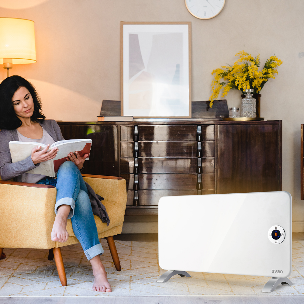 Mujer lee un libro cerca de un calefactor eléctrico blanco. Silla amarilla, armario y planta en el fondo.