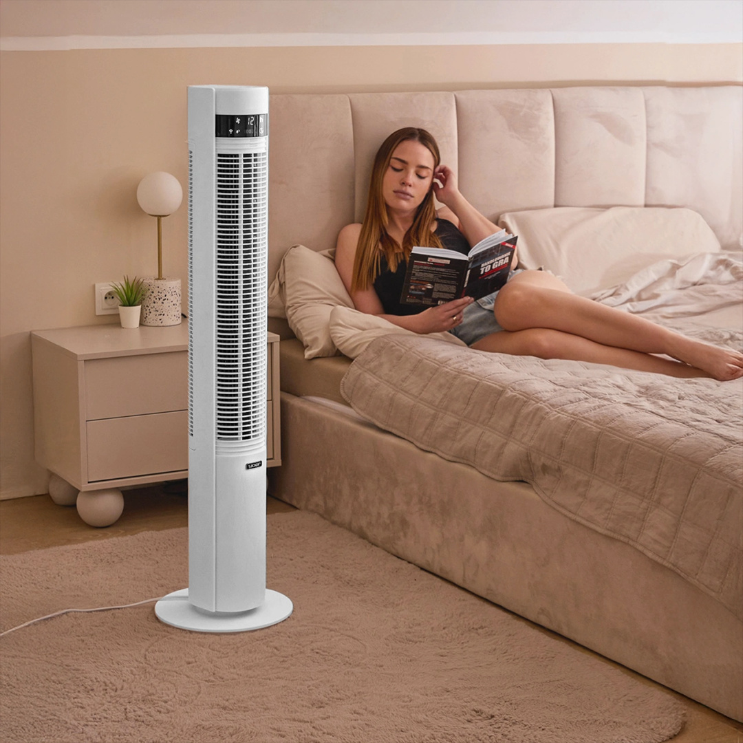 Kobieta czyta książkę w łóżku z białym wentylatorem wieżowym w tle.